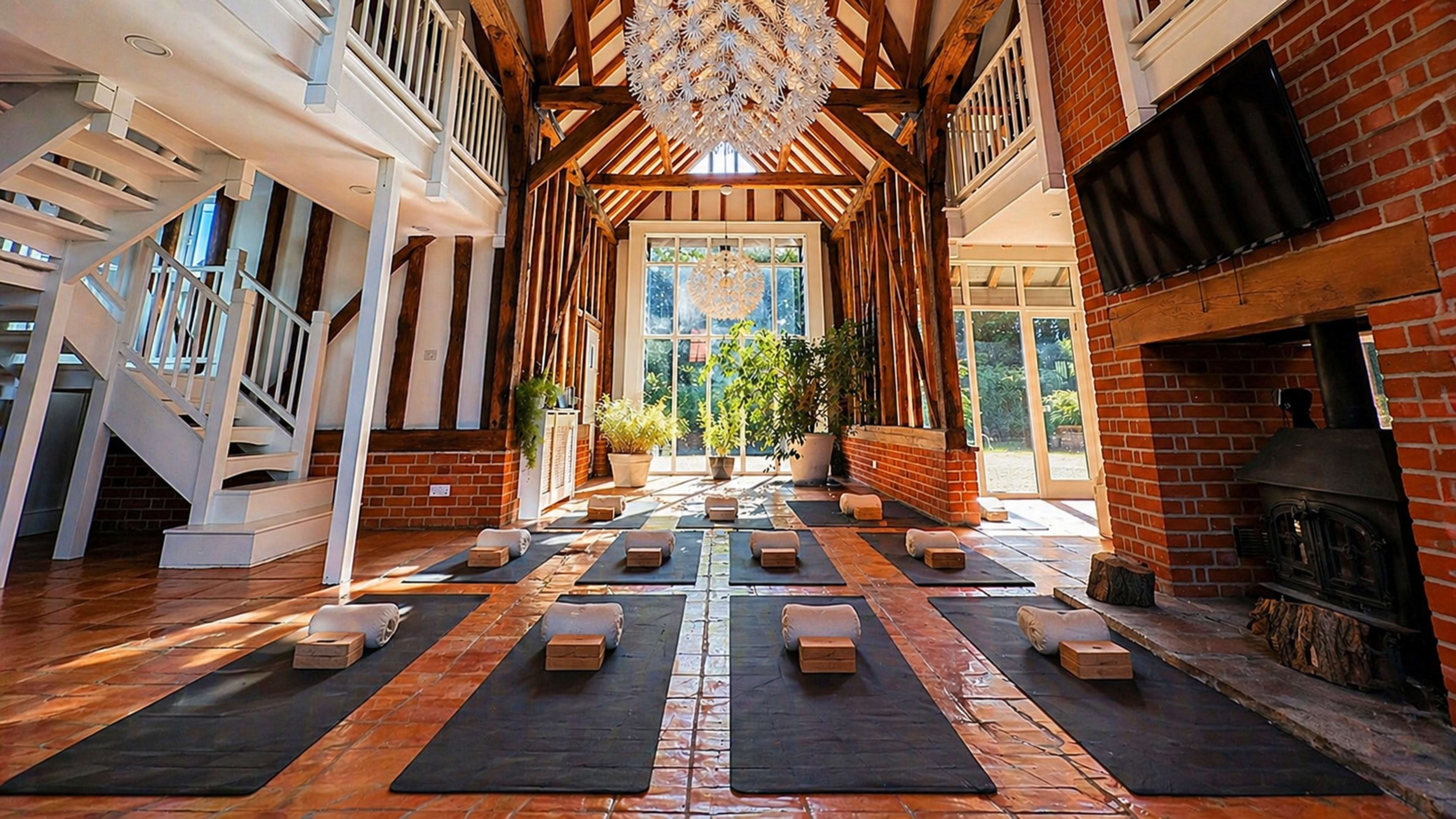 The image shows a spacious, rustic yoga studio with wooden beams, yoga mats arranged neatly on the floor, and a large window allowing natural light to enter.