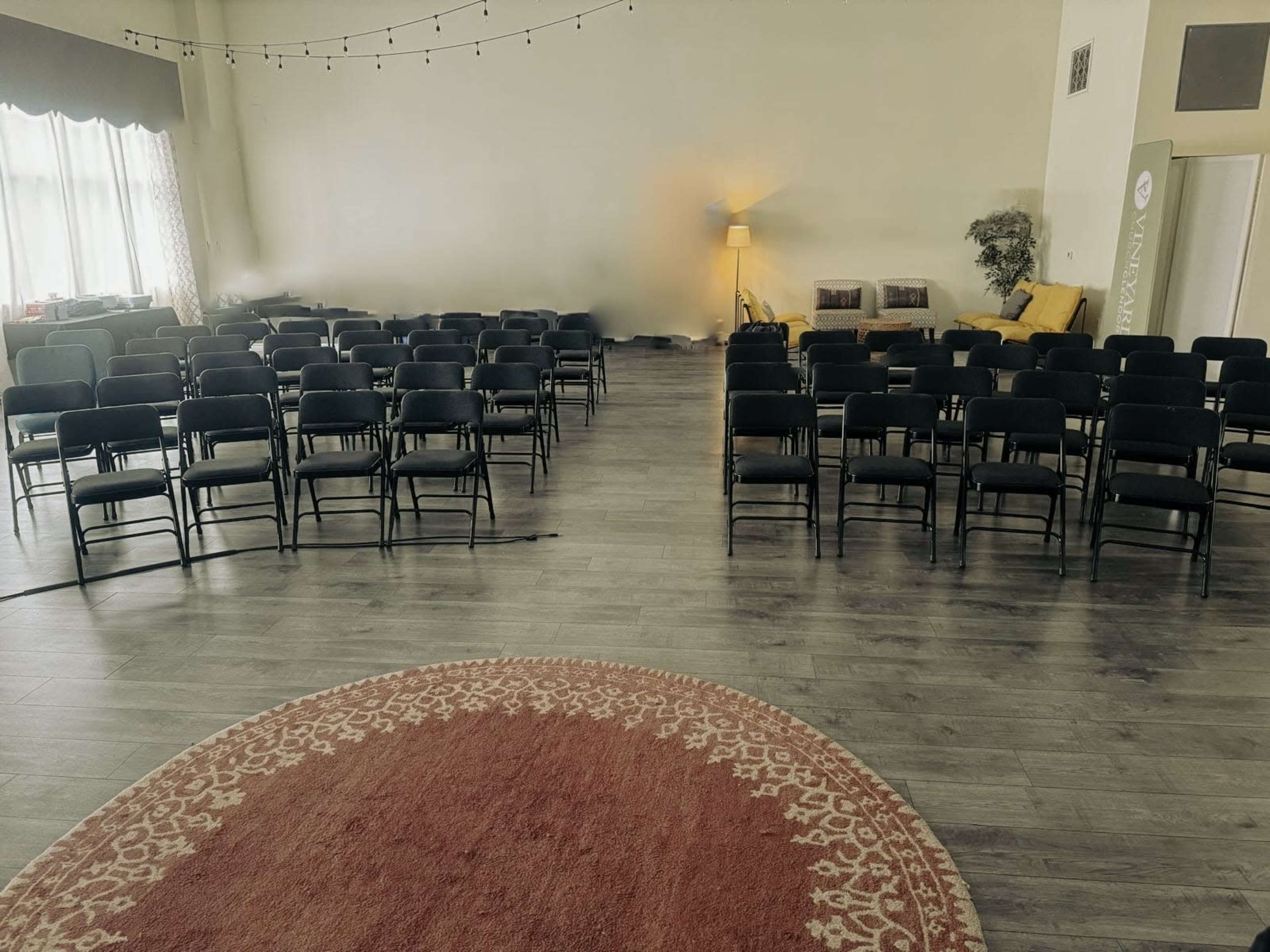 The image shows a spacious room arranged with rows of black chairs facing a central area, along with a circular rug, a sofa, and a lamp in the background.