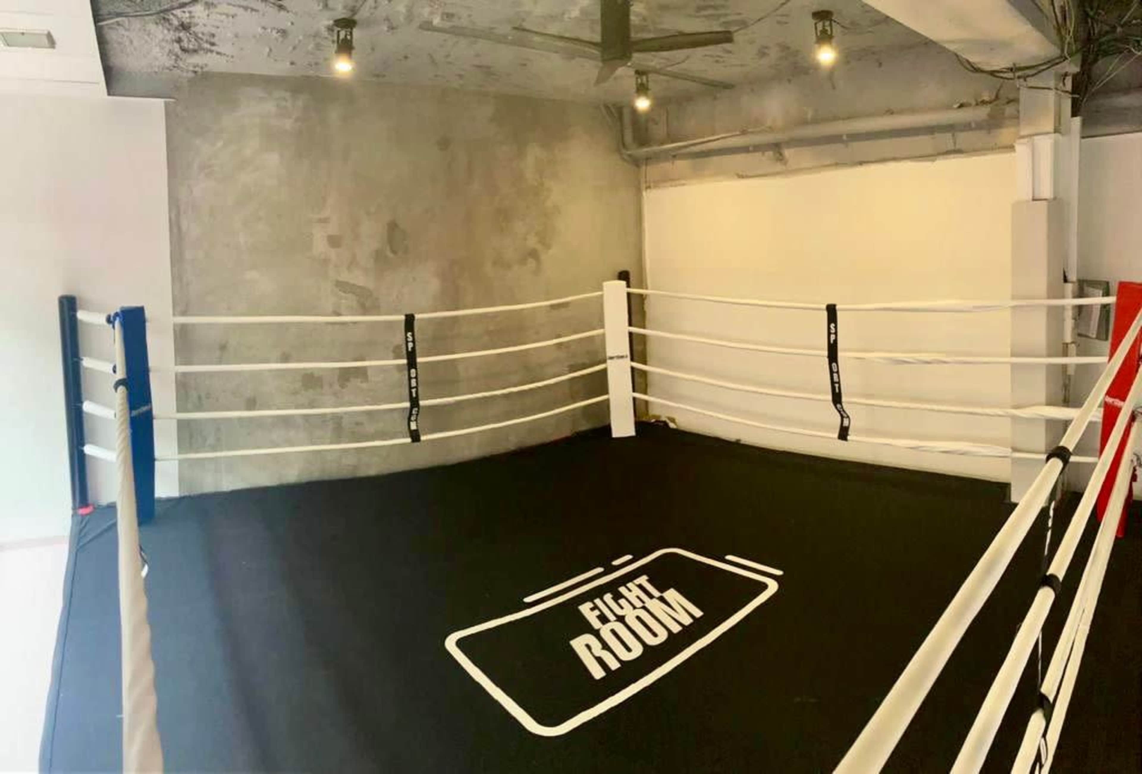 A boxing ring with white ropes and a black floor mat labeled "FIGHT ROOM" situated in a minimalist room with exposed concrete walls.