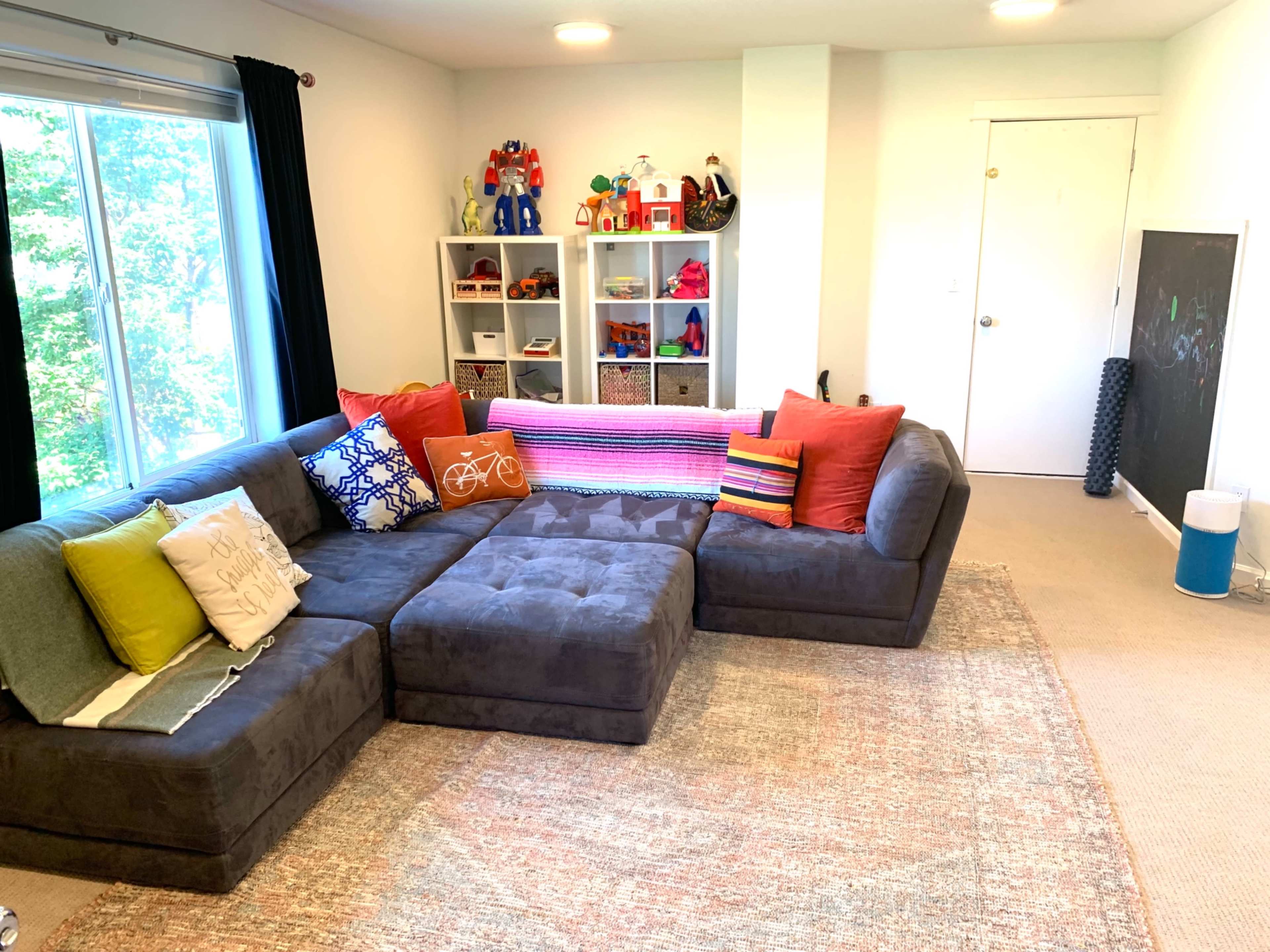 A modern living room features a large gray sectional couch with colorful pillows, a light rug, and a bookshelf filled with toys and children's items.