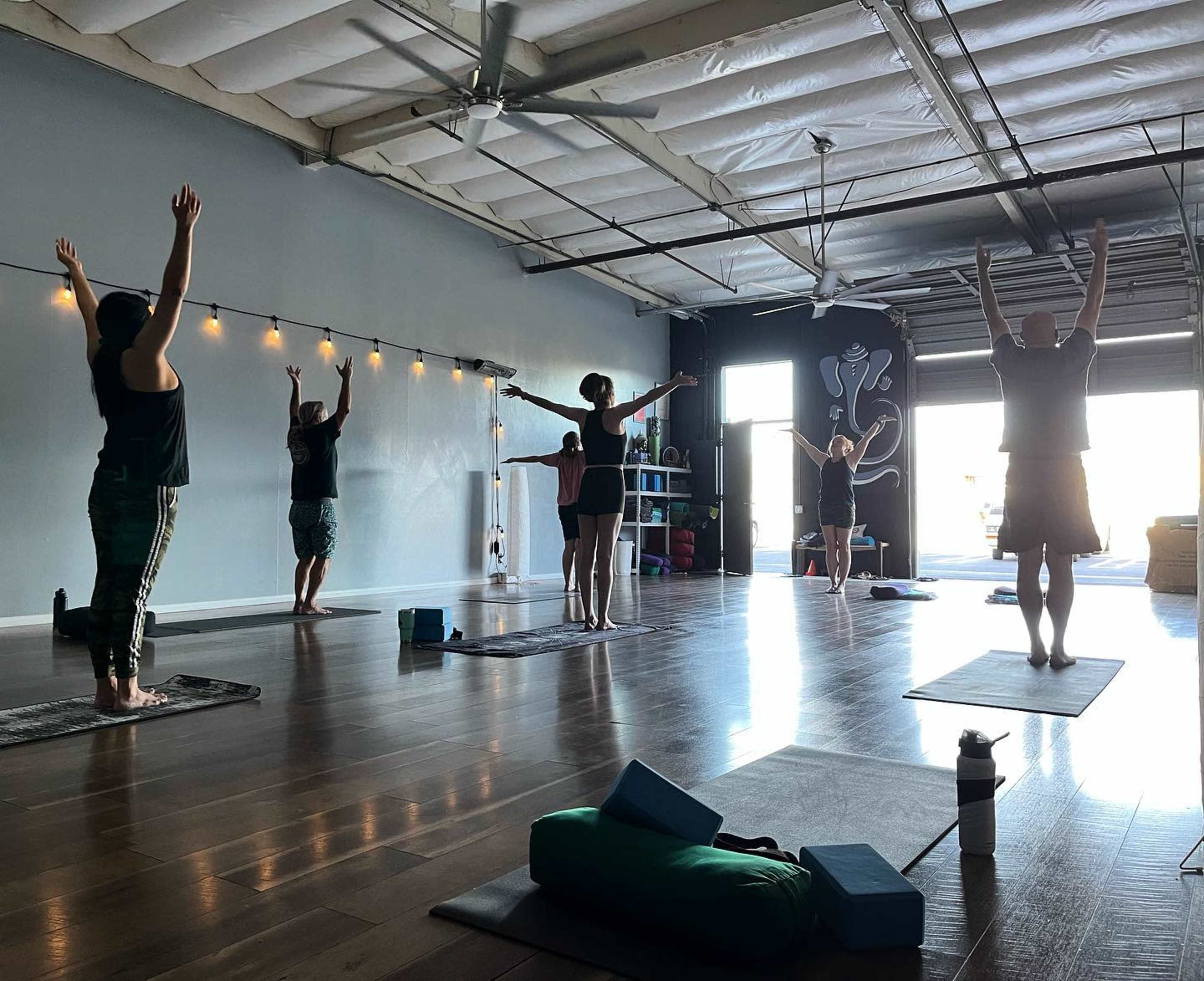 A group of six people is practicing yoga in a spacious studio with wooden flooring and large windows.