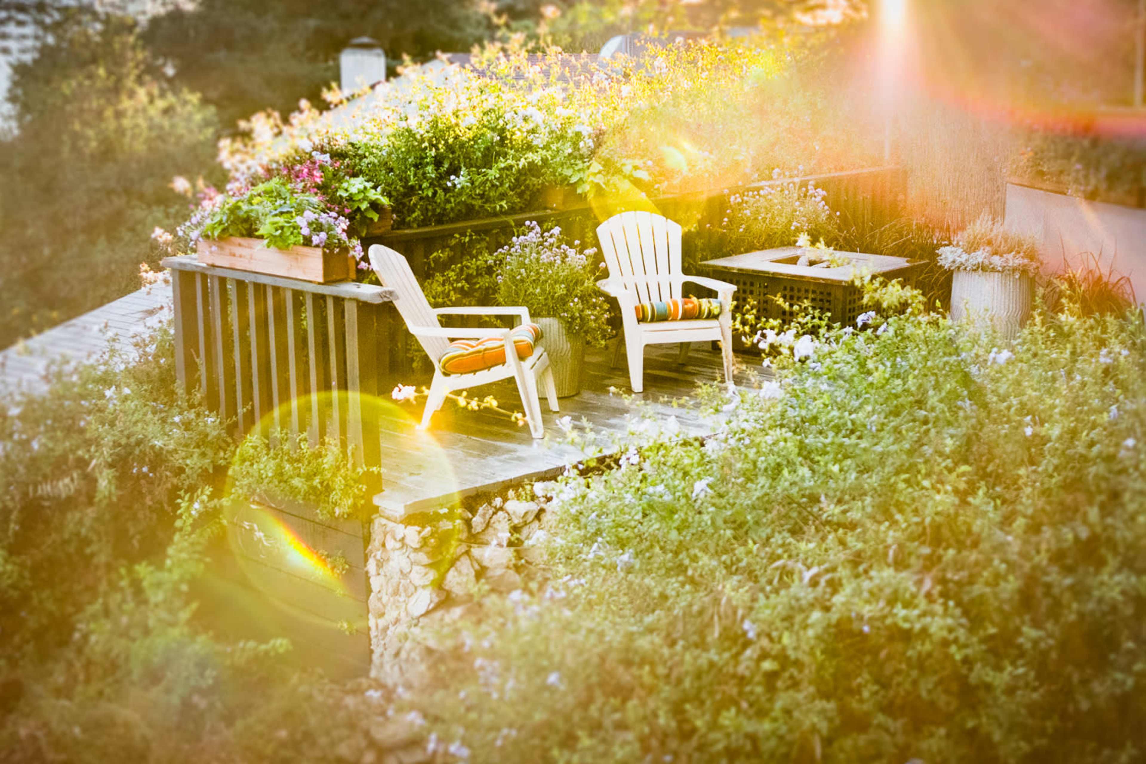 A wooden deck surrounded by colorful flowers features two white plastic chairs and a small table.