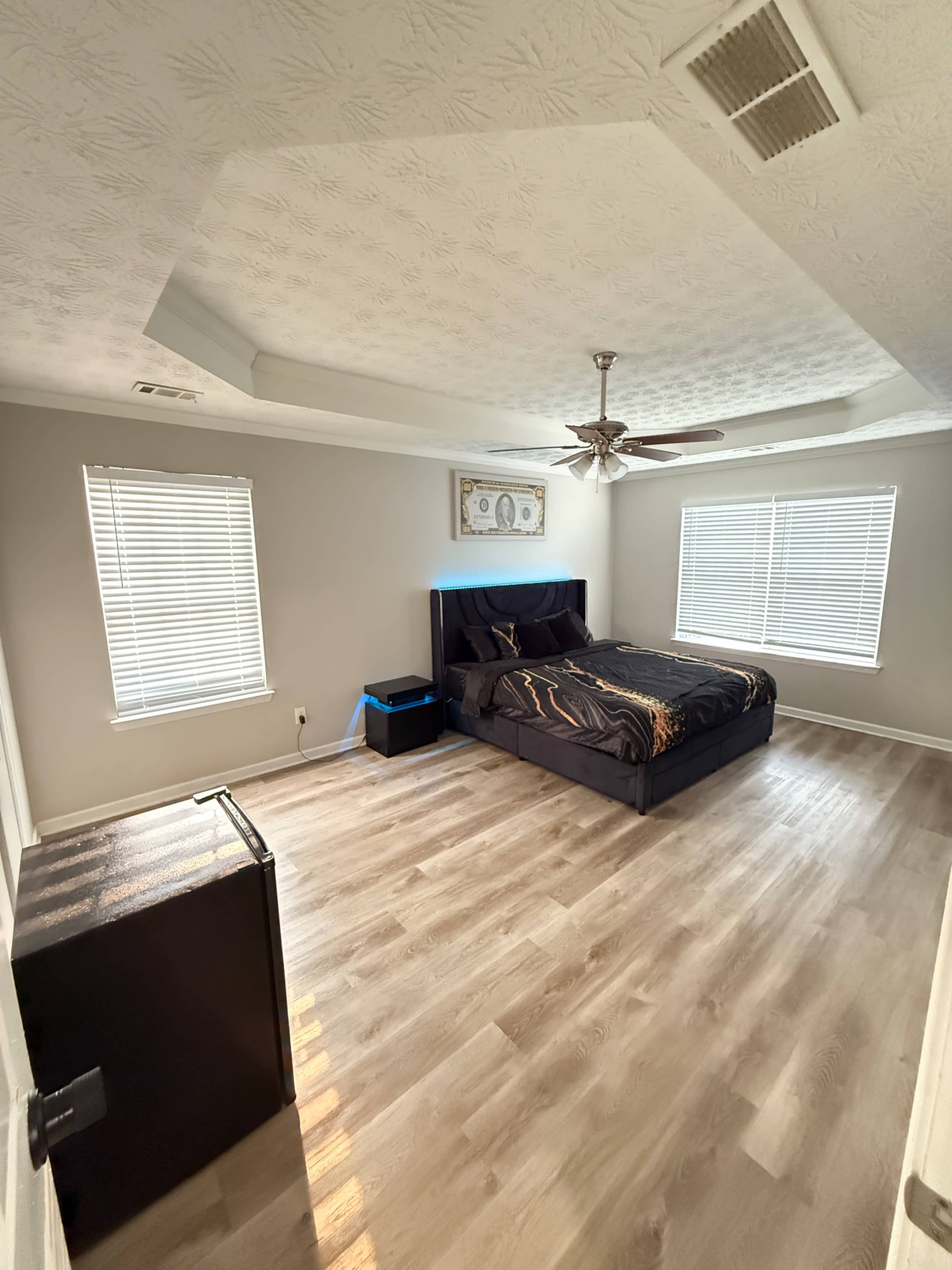 The image shows a spacious bedroom featuring a black bed with a patterned comforter, large windows with blinds, and a small black nightstand.