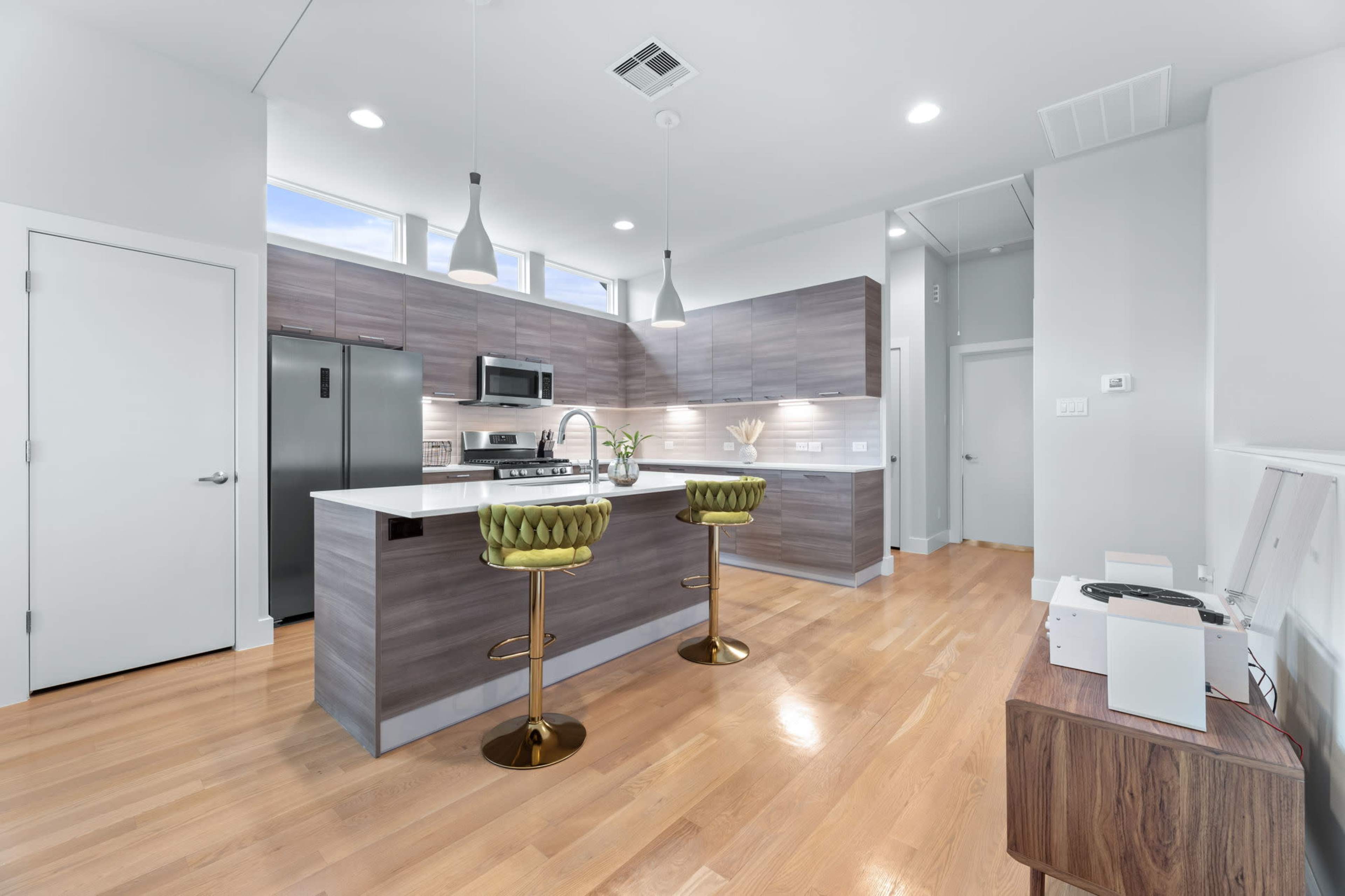 A modern kitchen features a central island with two green bar stools, stainless steel appliances, and light hardwood flooring.