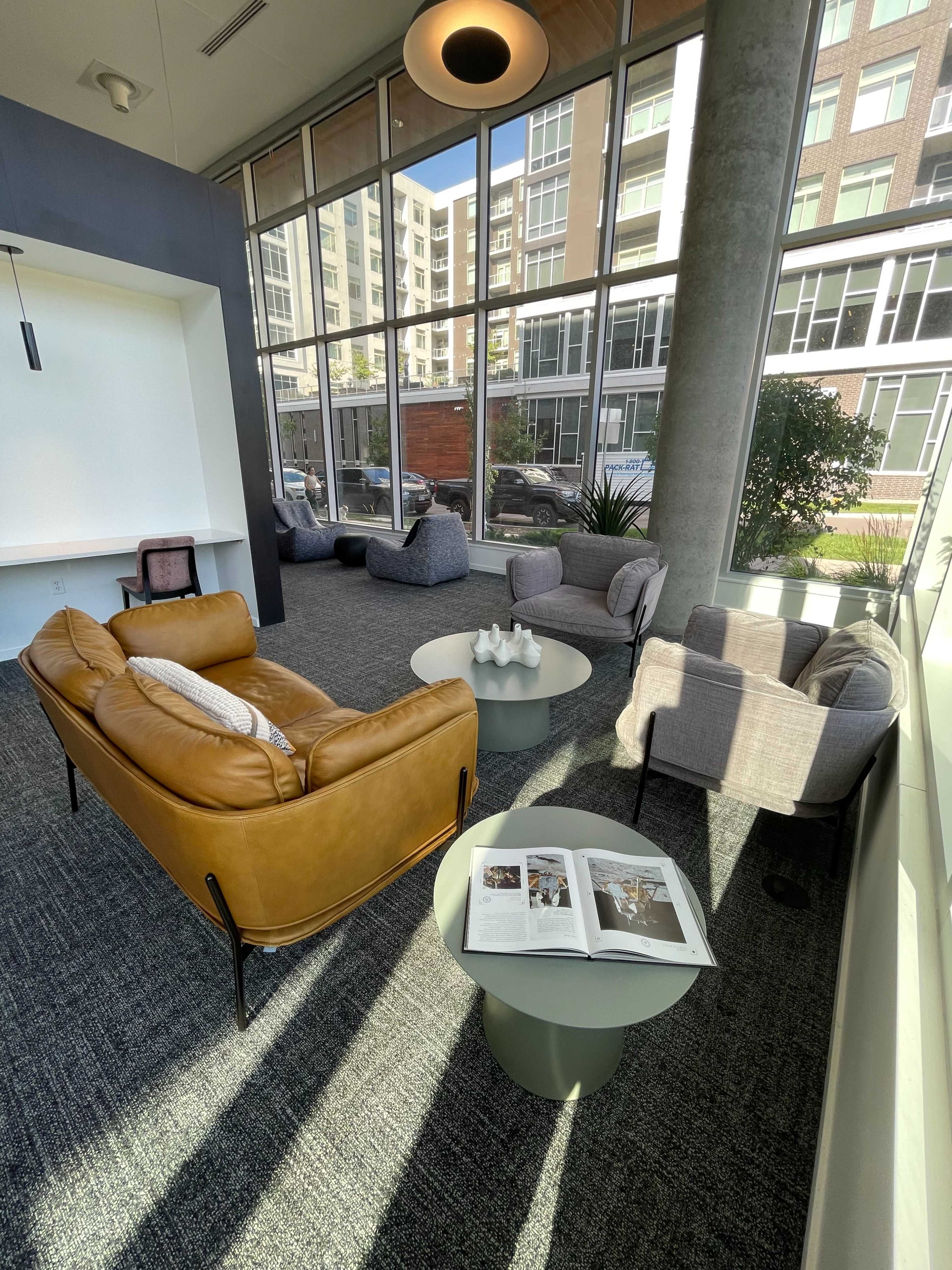 A modern lounge area with a brown leather sofa, light gray armchairs, a round coffee table, and large windows allowing natural light to illuminate the space.