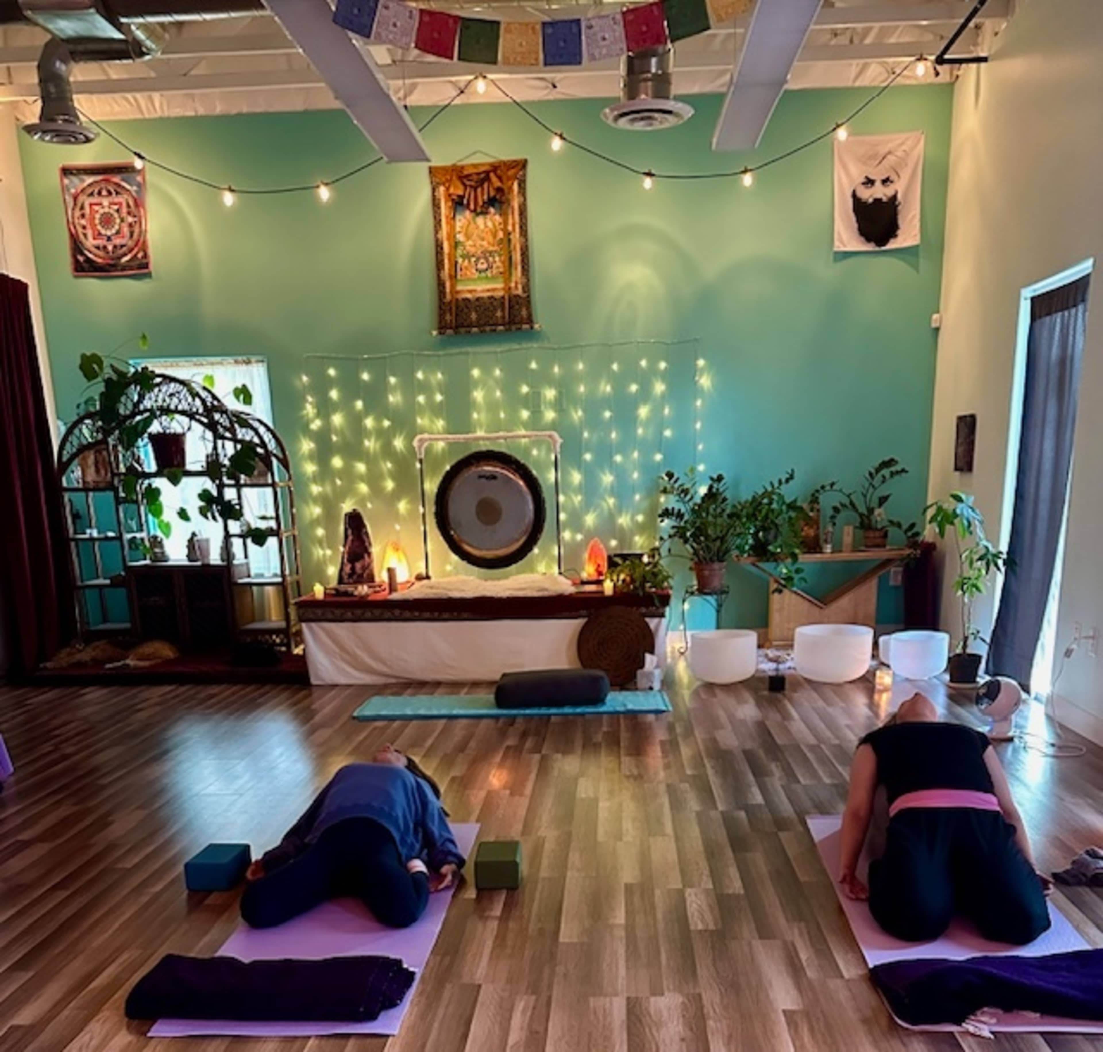 The image shows a serene yoga studio with two individuals practicing a forward bend pose on yoga mats, surrounded by plants, decorative wall hangings, and soft lighting.