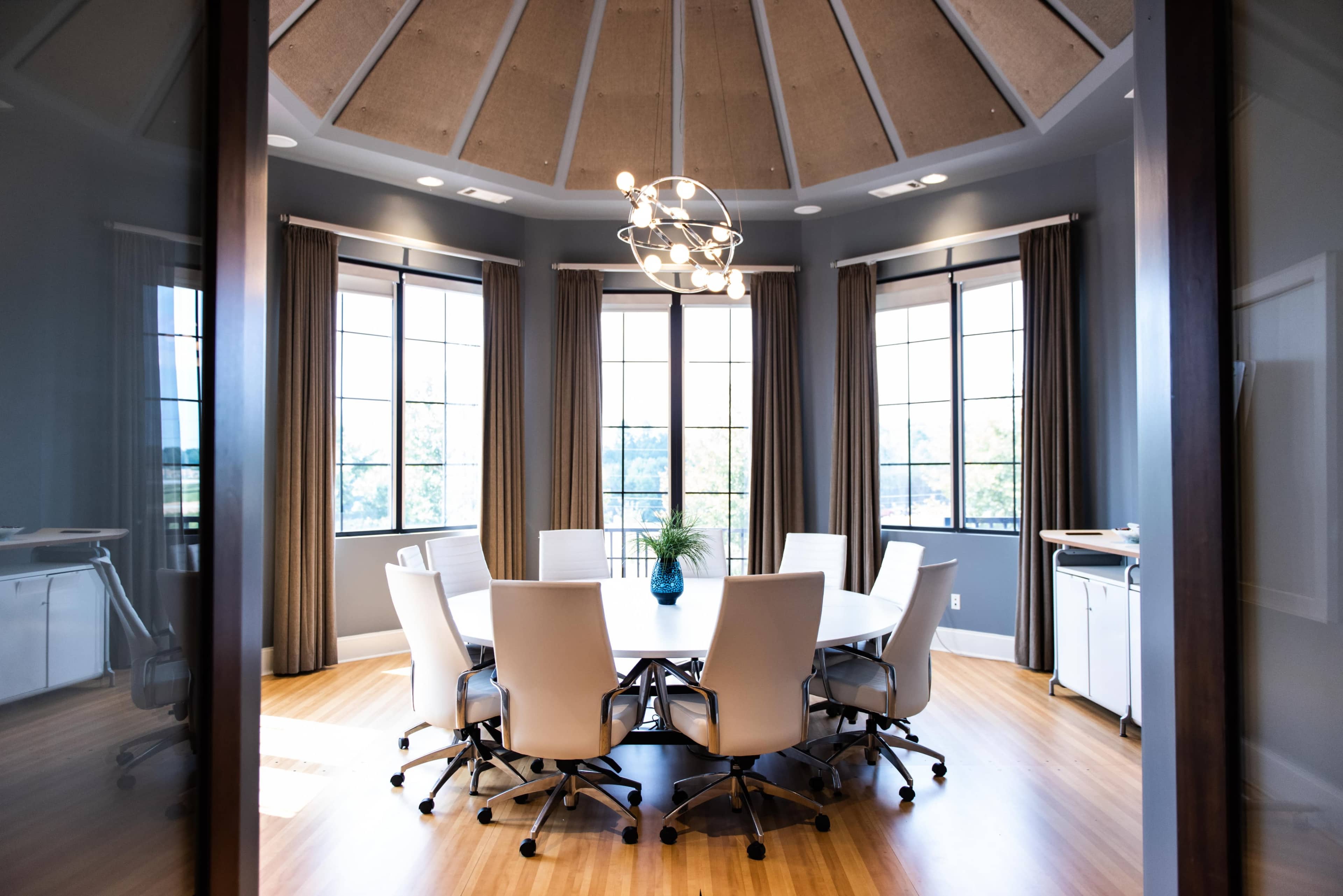 A modern conference room features a round table surrounded by white chairs, large windows, and a central light fixture.