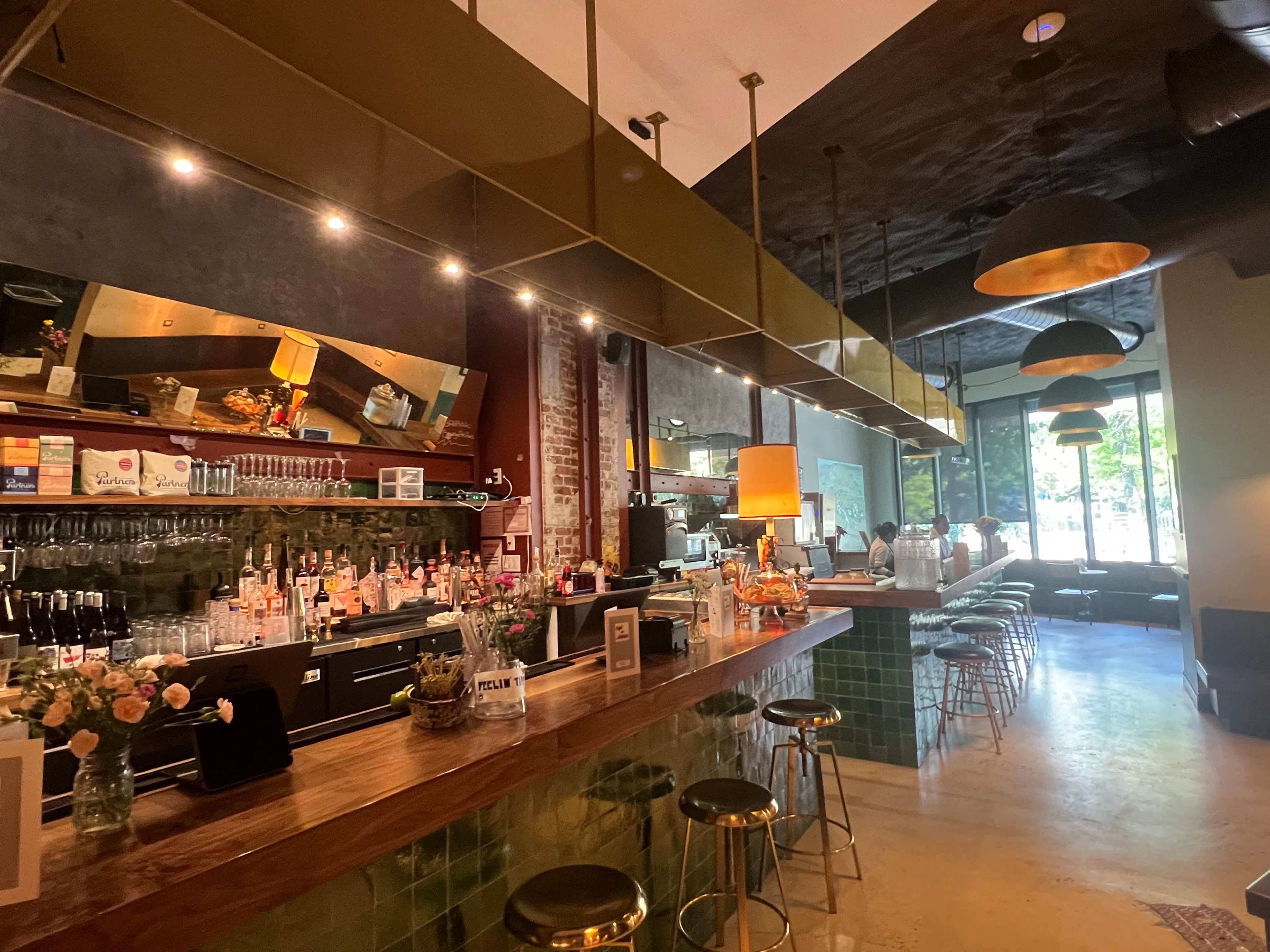 The image shows a modern bar interior with a wooden counter, high stools, and shelves displaying various bottles, illuminated by pendant lights.