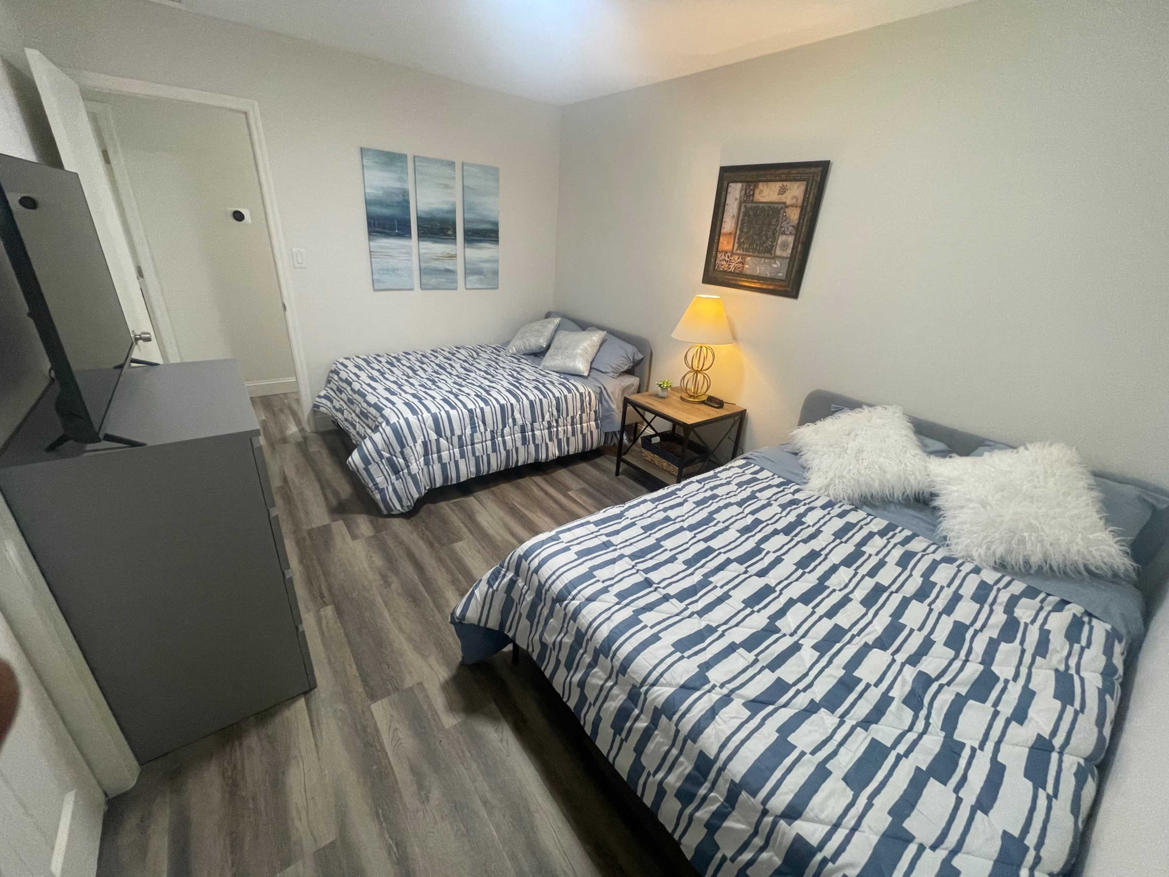 A well-arranged bedroom with two beds, a television, and light-colored walls, featuring ocean-themed artwork and a lamp on a bedside table.