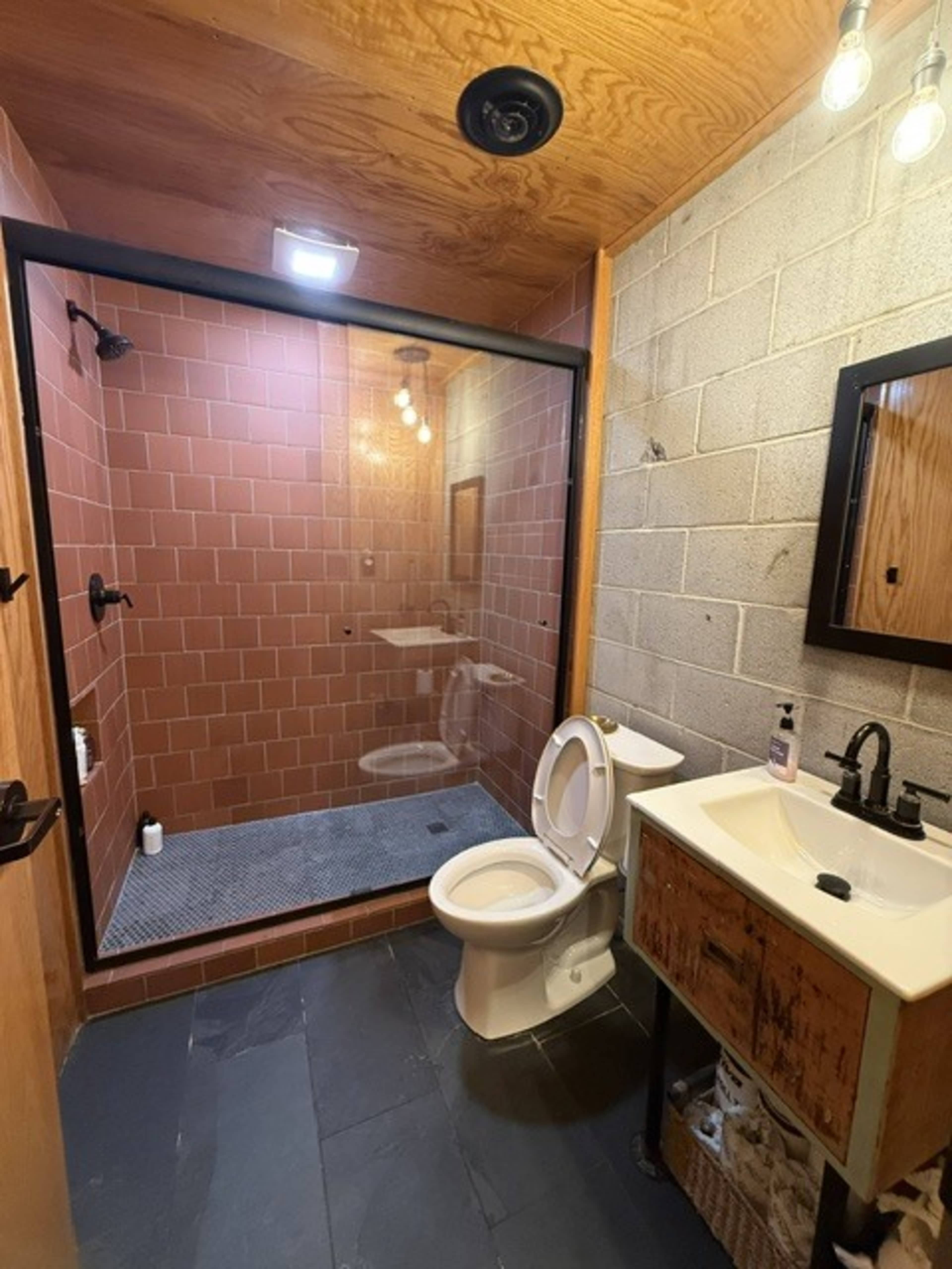 The image shows a modern bathroom featuring a glass-enclosed shower, a toilet, and a sink with a wooden cabinet, all set against a backdrop of textured walls and wooden ceiling.