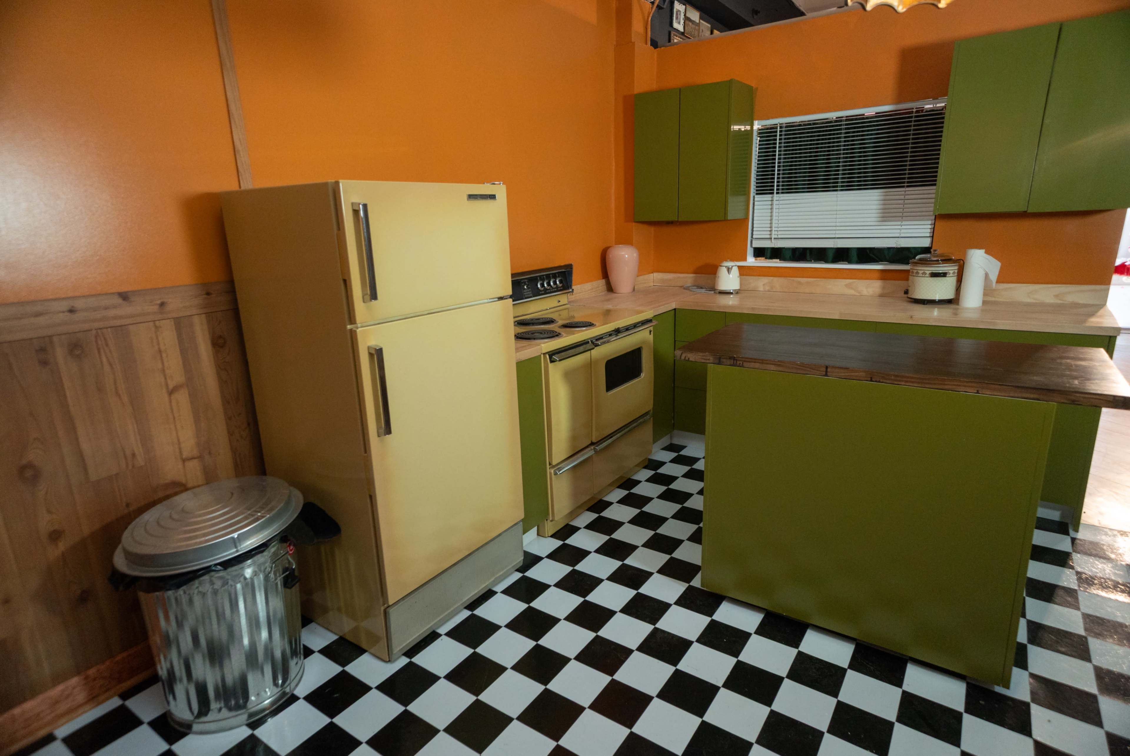 The image shows a retro kitchen with orange walls, green cabinets, a beige refrigerator, and a black and white checkered floor.