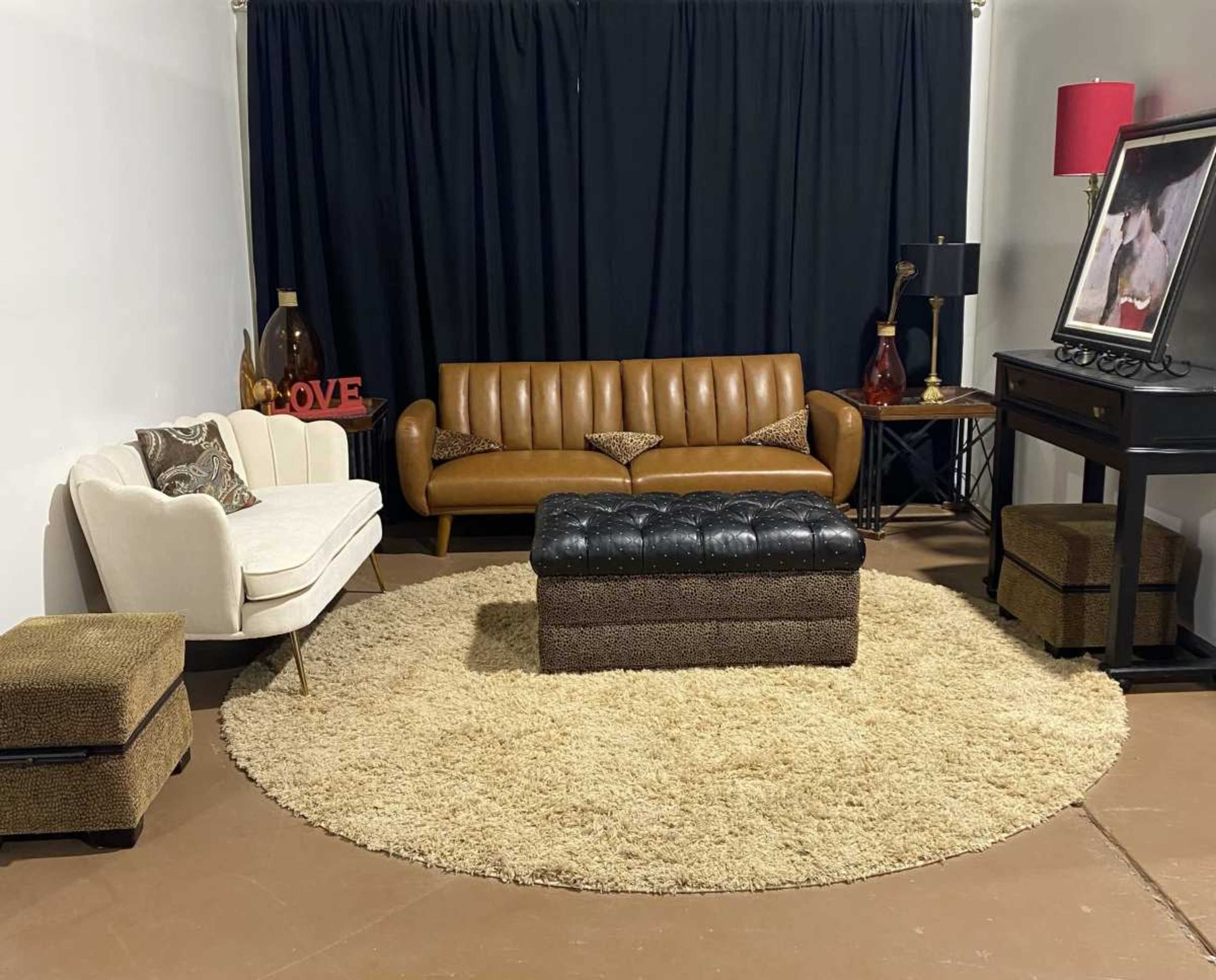 The scene features a stylish living room with a brown leather sofa, a white armchair, a round area rug, a black coffee table, and various decorative elements against a black curtain backdrop.