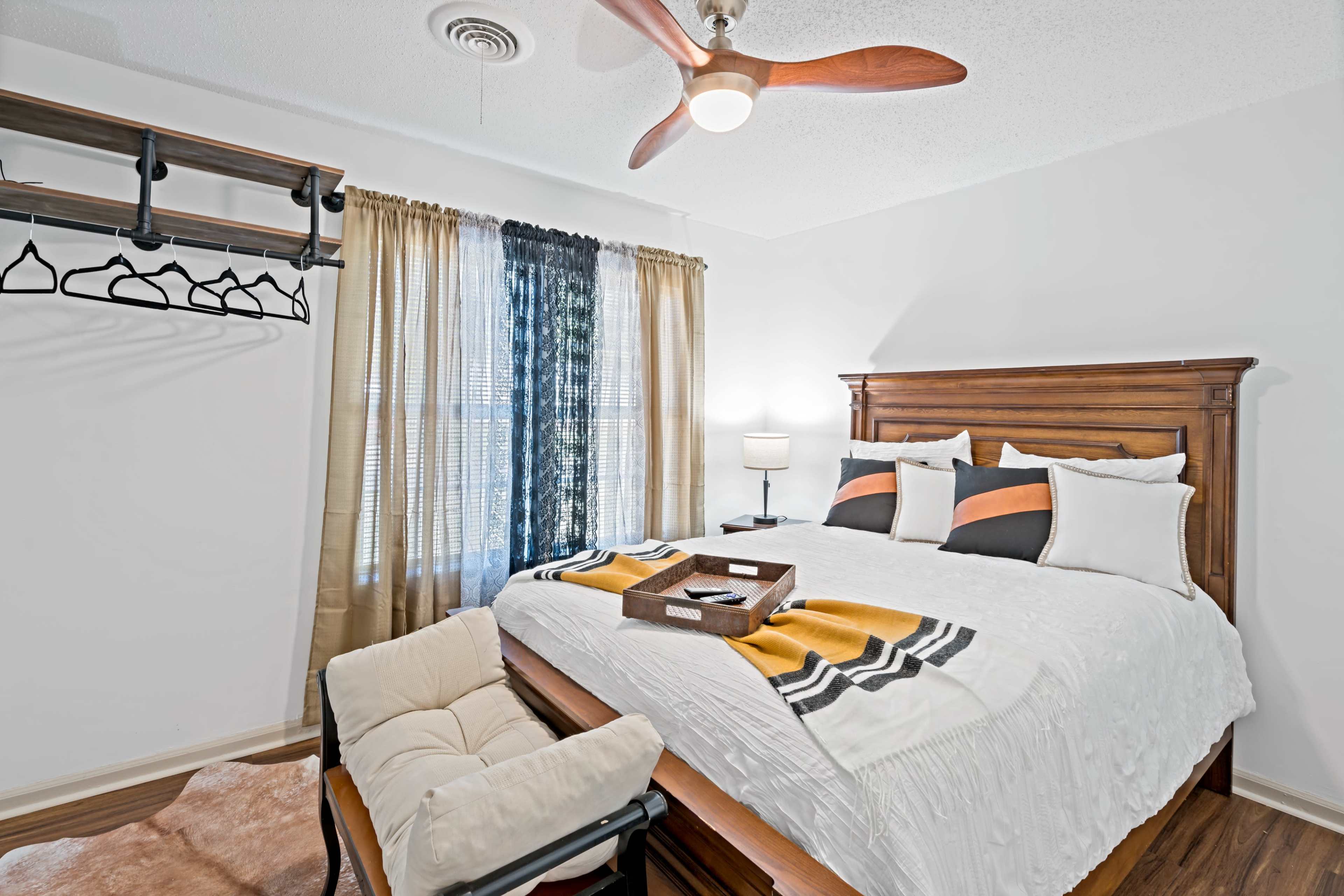 The image shows a neatly arranged bedroom featuring a wooden bed with white bedding, a small seating area, and a wall-mounted coat rack.