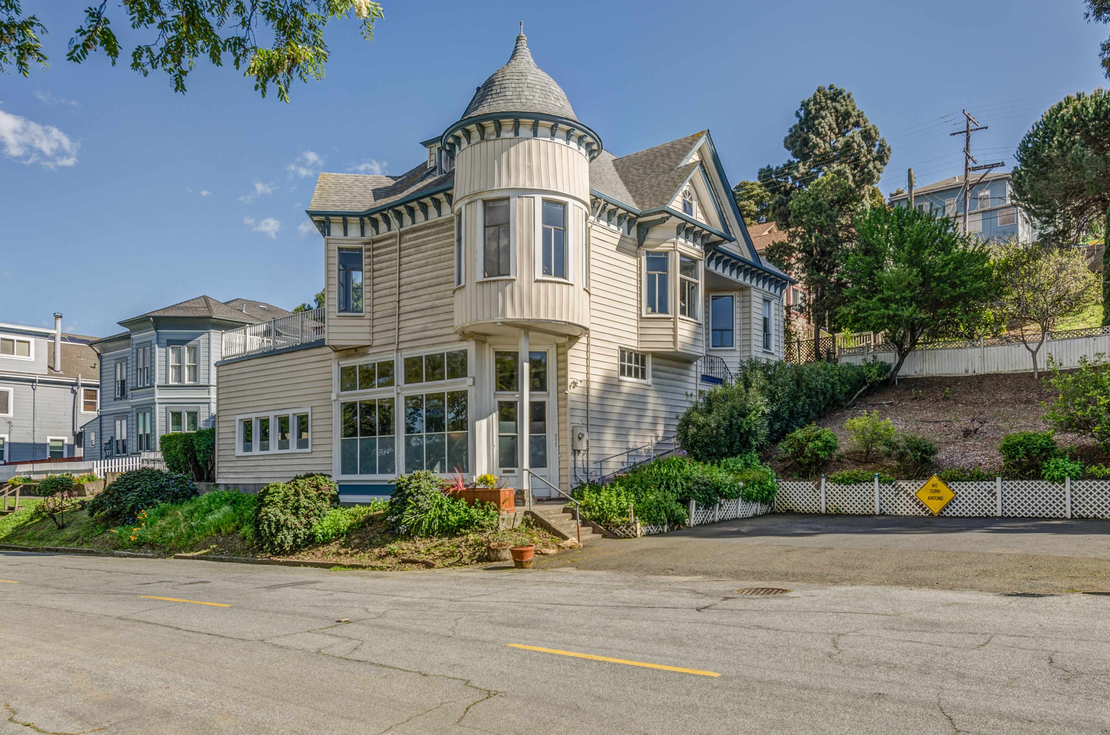 A historic Victorian-style house with a turret is situated on a tree-lined street.