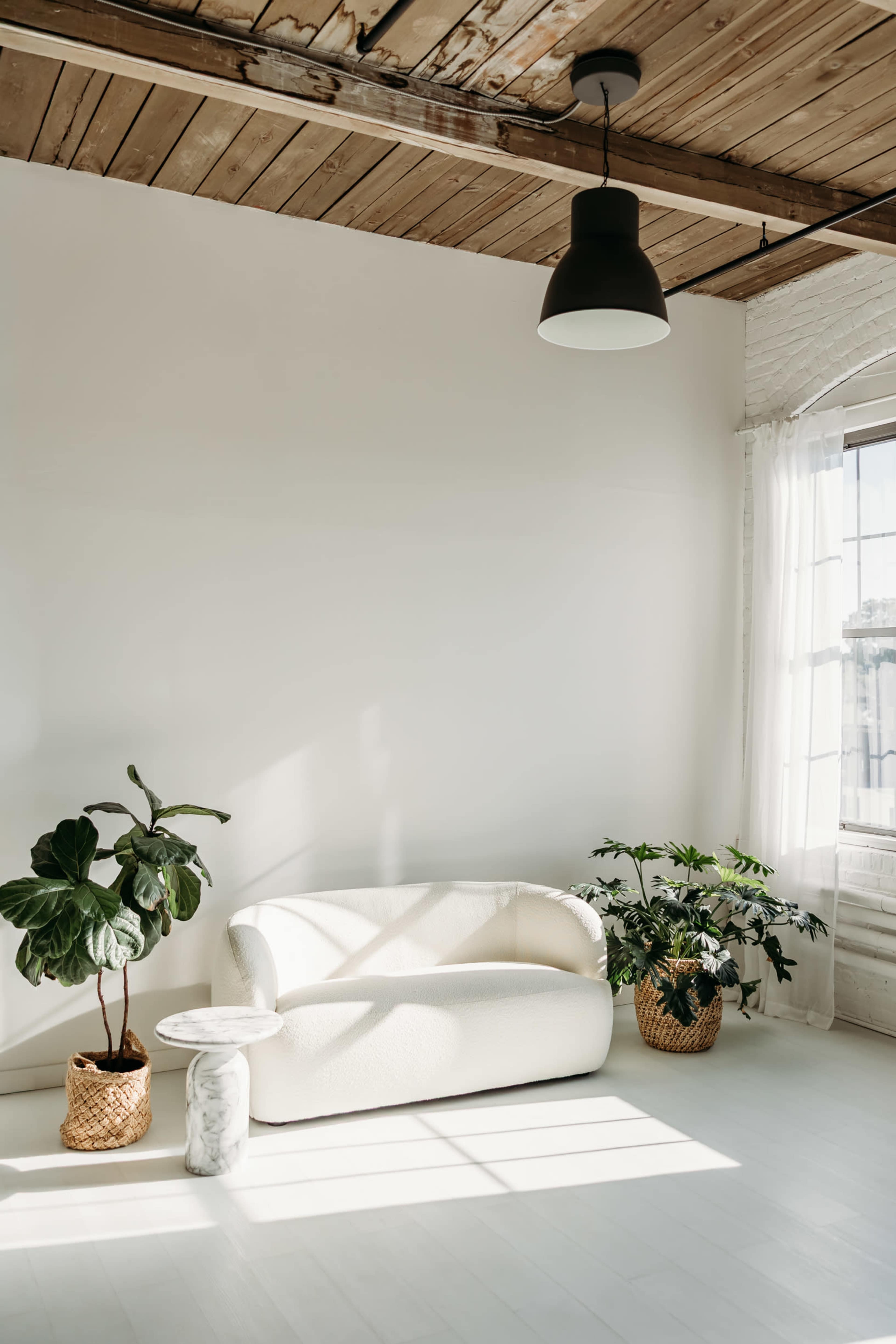The image shows a bright, minimalist living space featuring a white curved sofa, two potted plants, a small white side table, and large windows with sheer curtains.