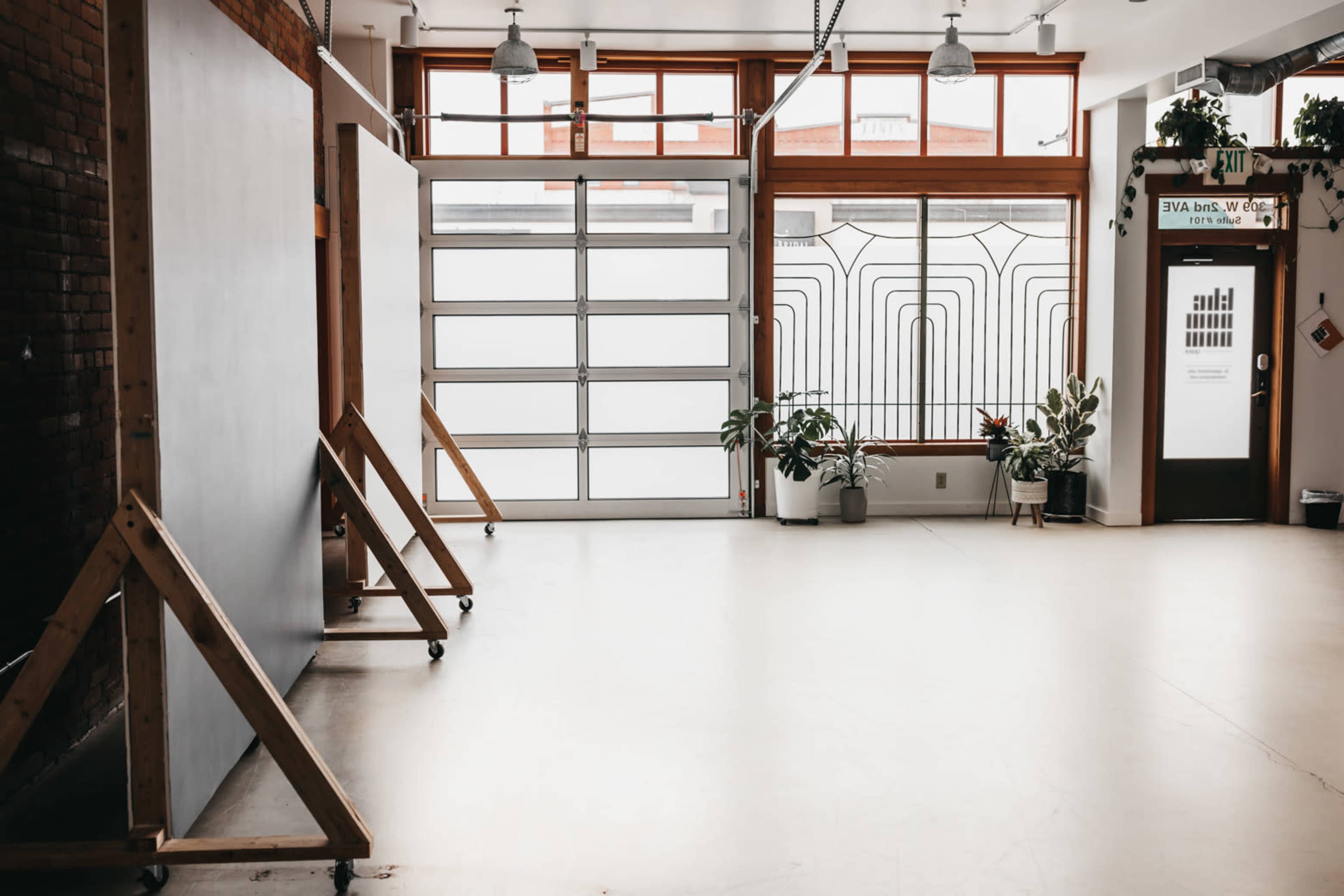A spacious, well-lit room with large windows, wooden frames standing on either side, and potted plants near an entrance door.