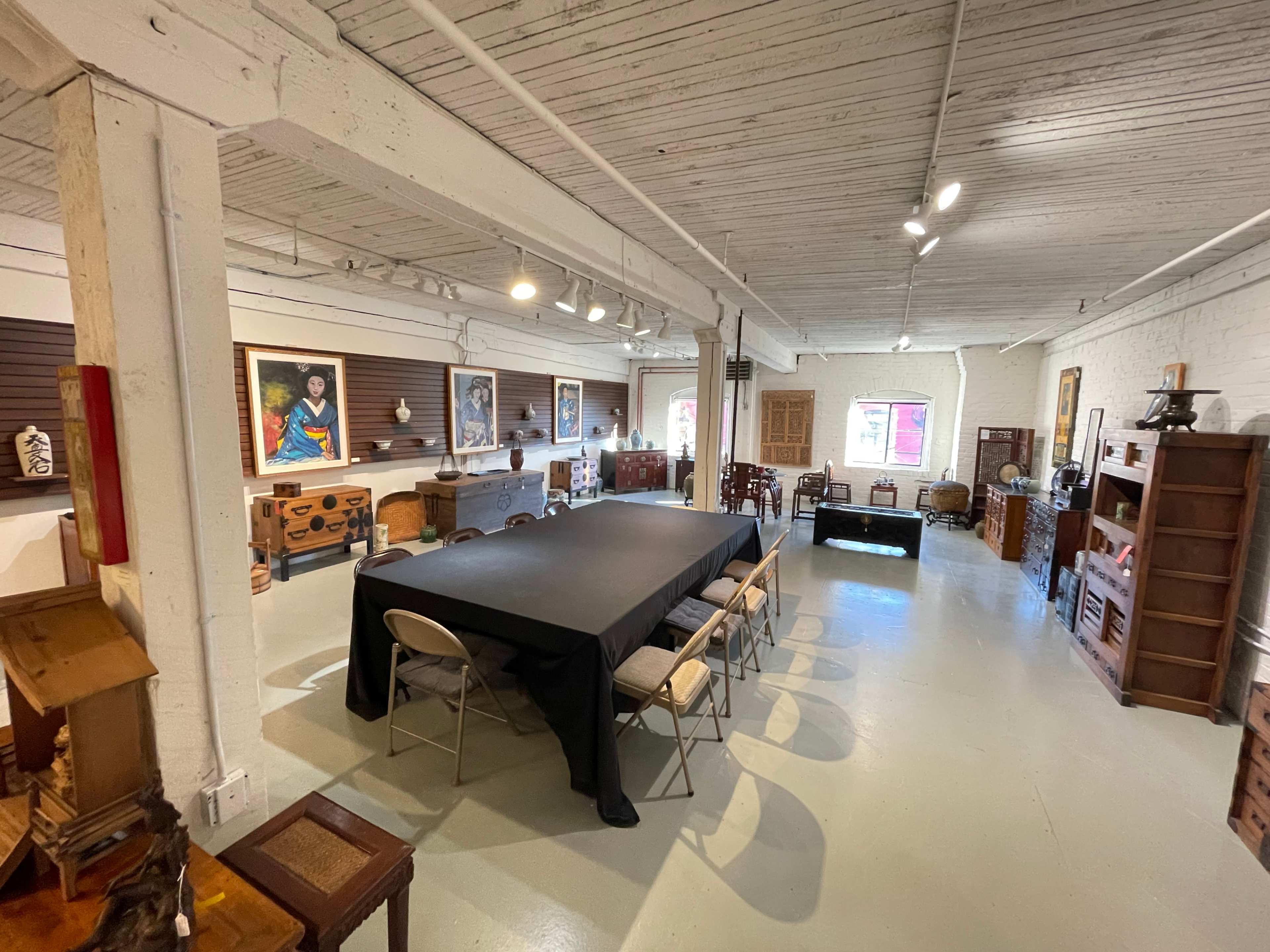 A spacious room with a large black table surrounded by chairs, artwork on the walls, and various wooden furniture pieces arranged throughout the space.
