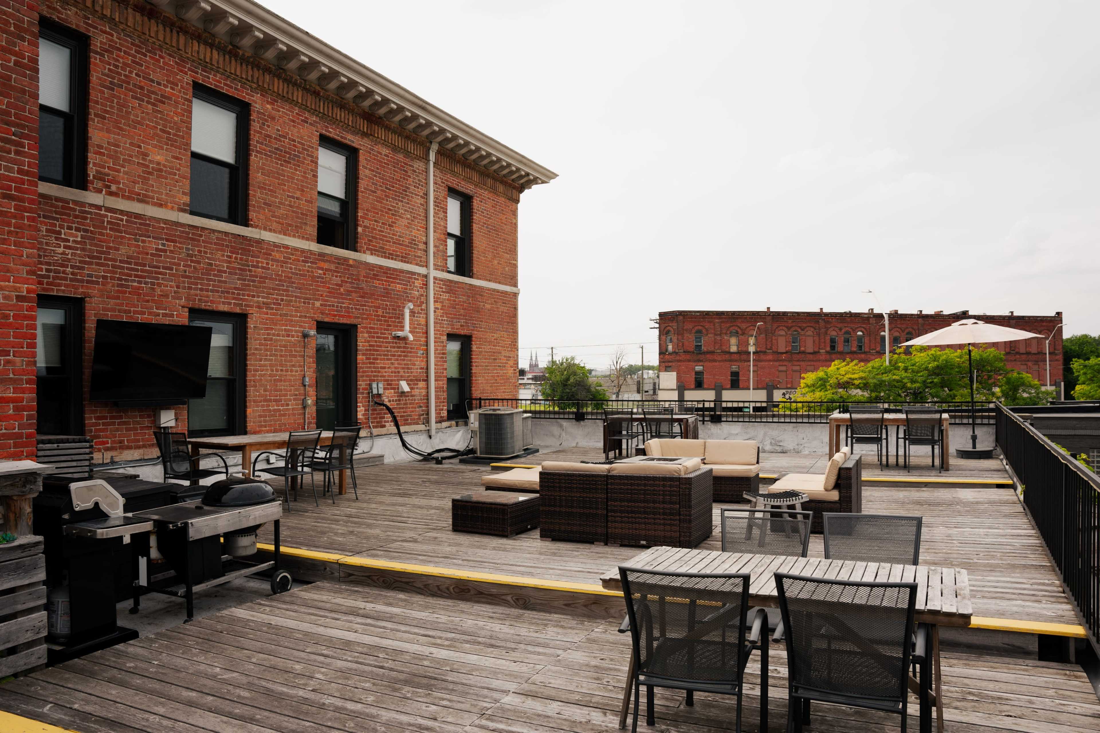A rooftop terrace with wooden flooring, outdoor furniture, and a view of a brick building in the background.