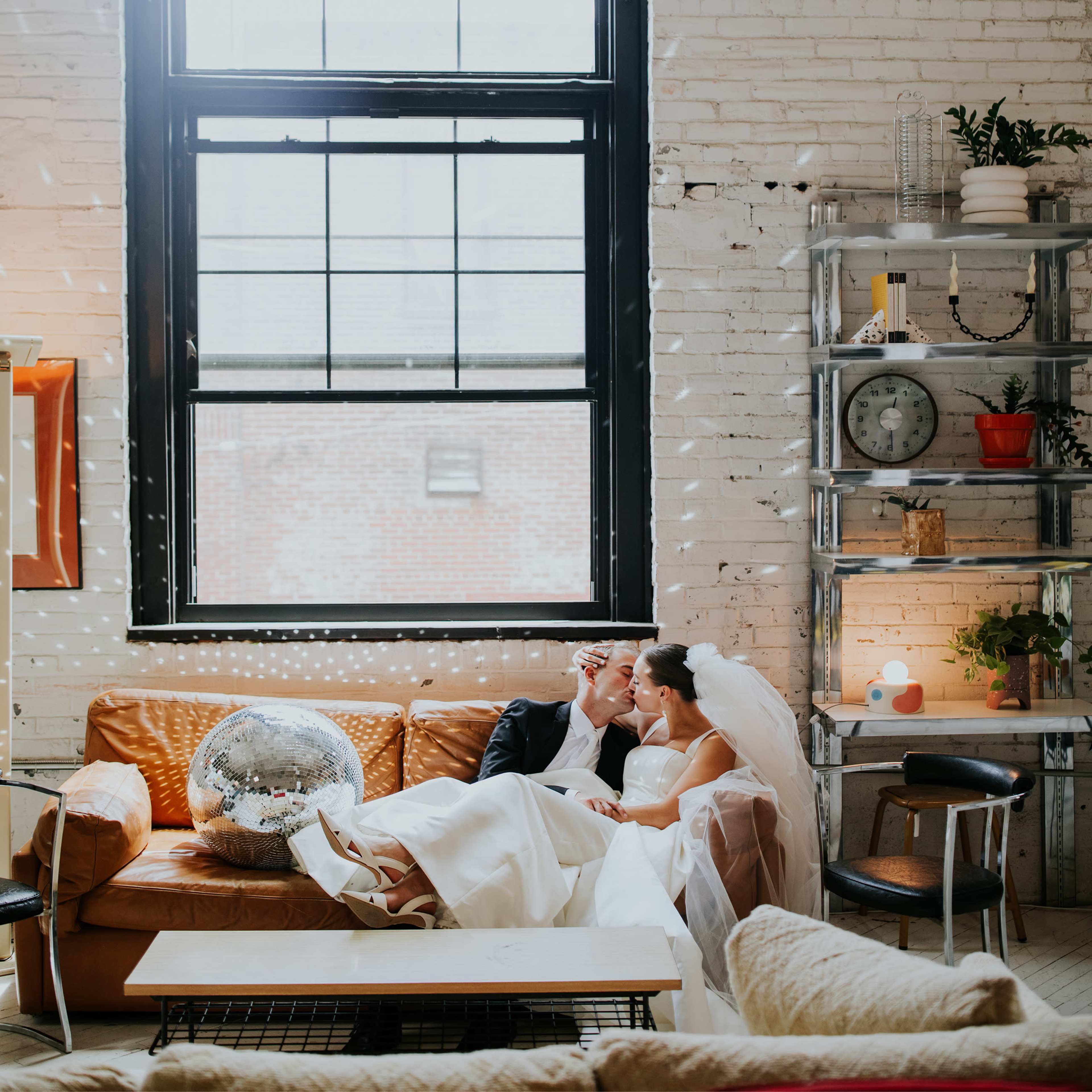 A bride and groom sit close together on a couch in a bright, industrial-style room decorated with a disco ball and various plants.