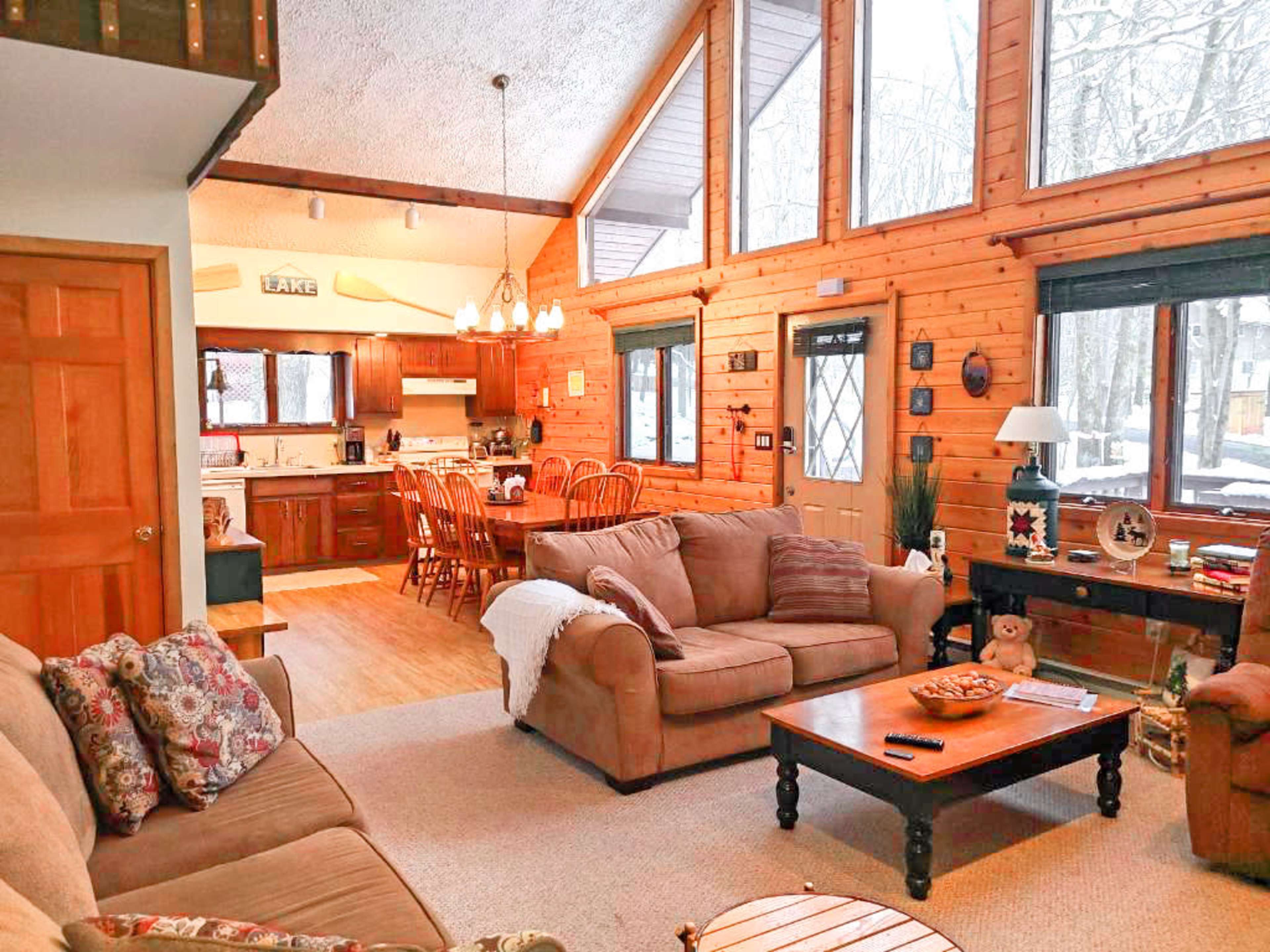 The image shows a cozy living area with wooden walls, large windows, a dining space, and a kitchen visible in the background.