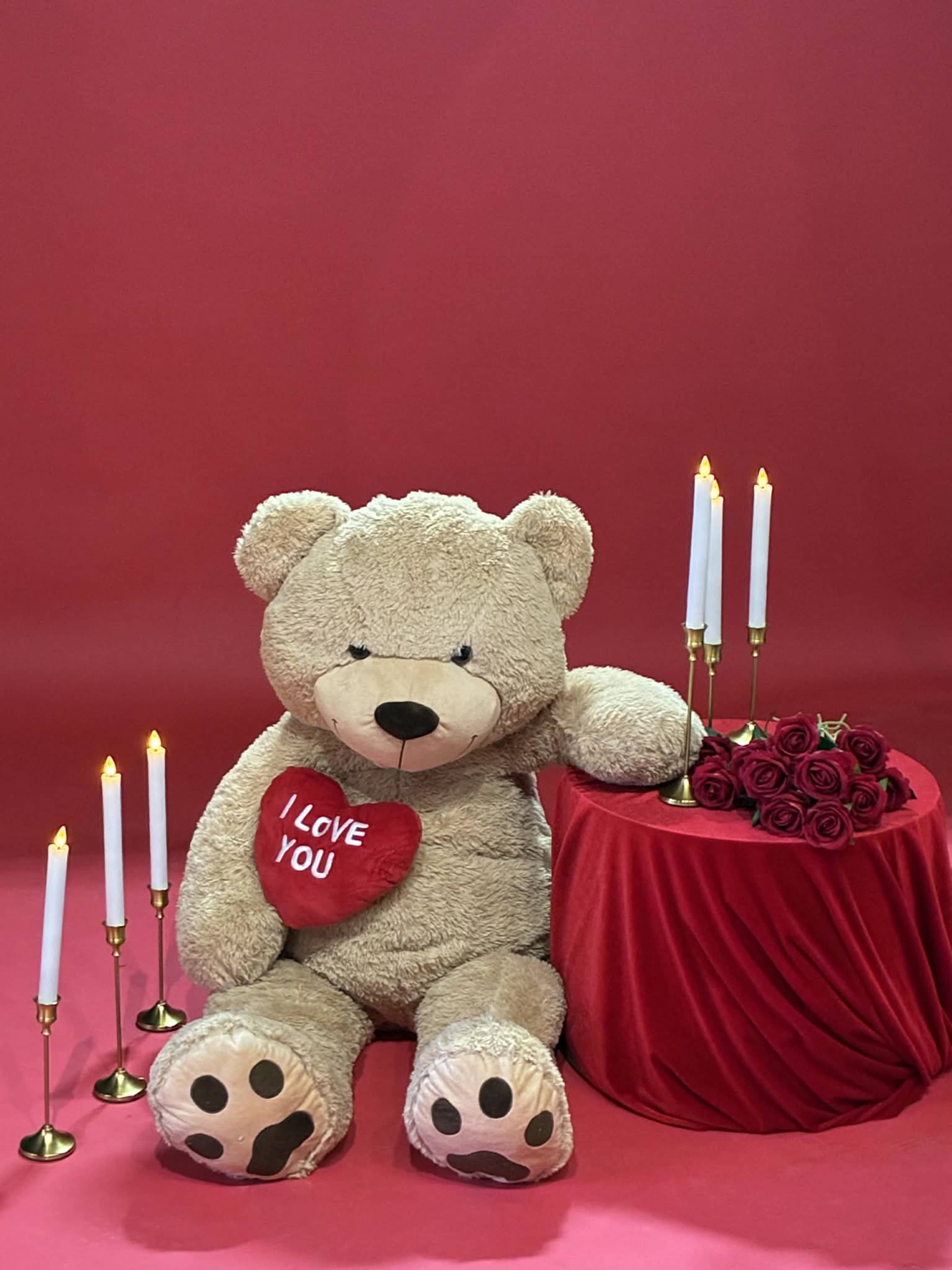 A large teddy bear holding a heart that says "I LOVE YOU" sits beside a table draped in red fabric with roses and lit candles against a red backdrop.