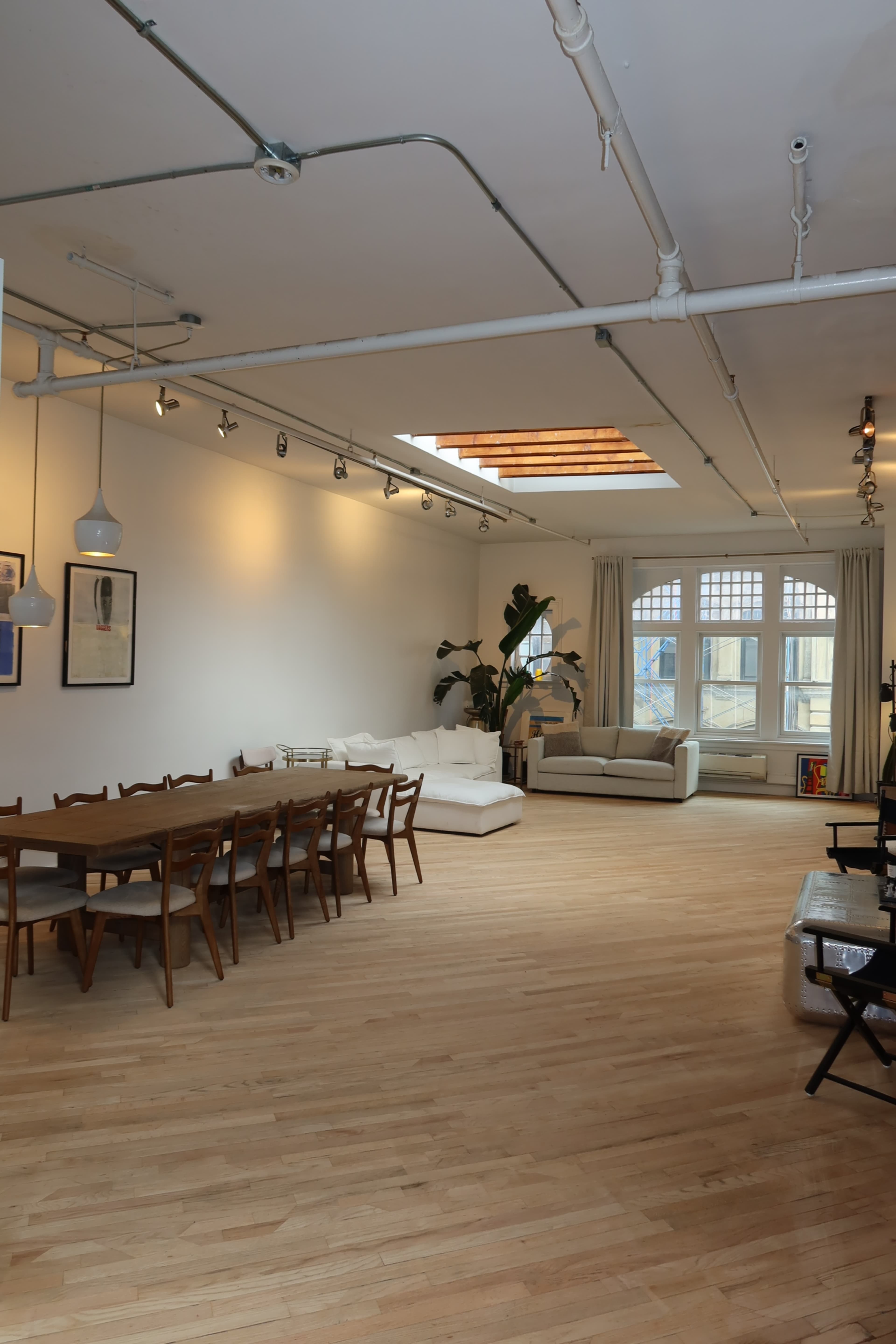 The image shows a spacious, well-lit interior of a modern living space featuring a long dining table, a white sofa, and large windows.
