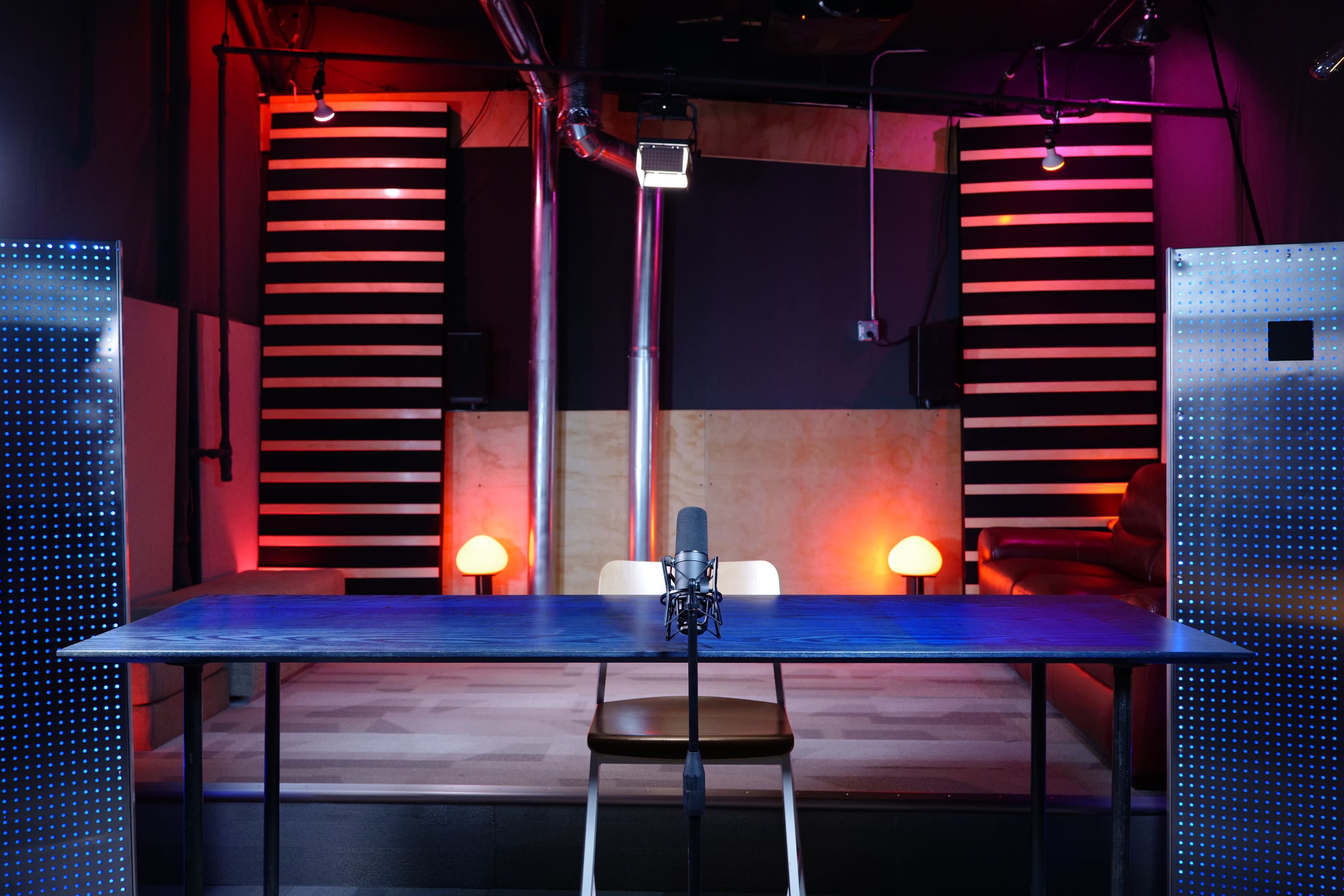 A well-lit studio with a microphone on a table facing a seating area, flanked by illuminated lamps and striped walls.