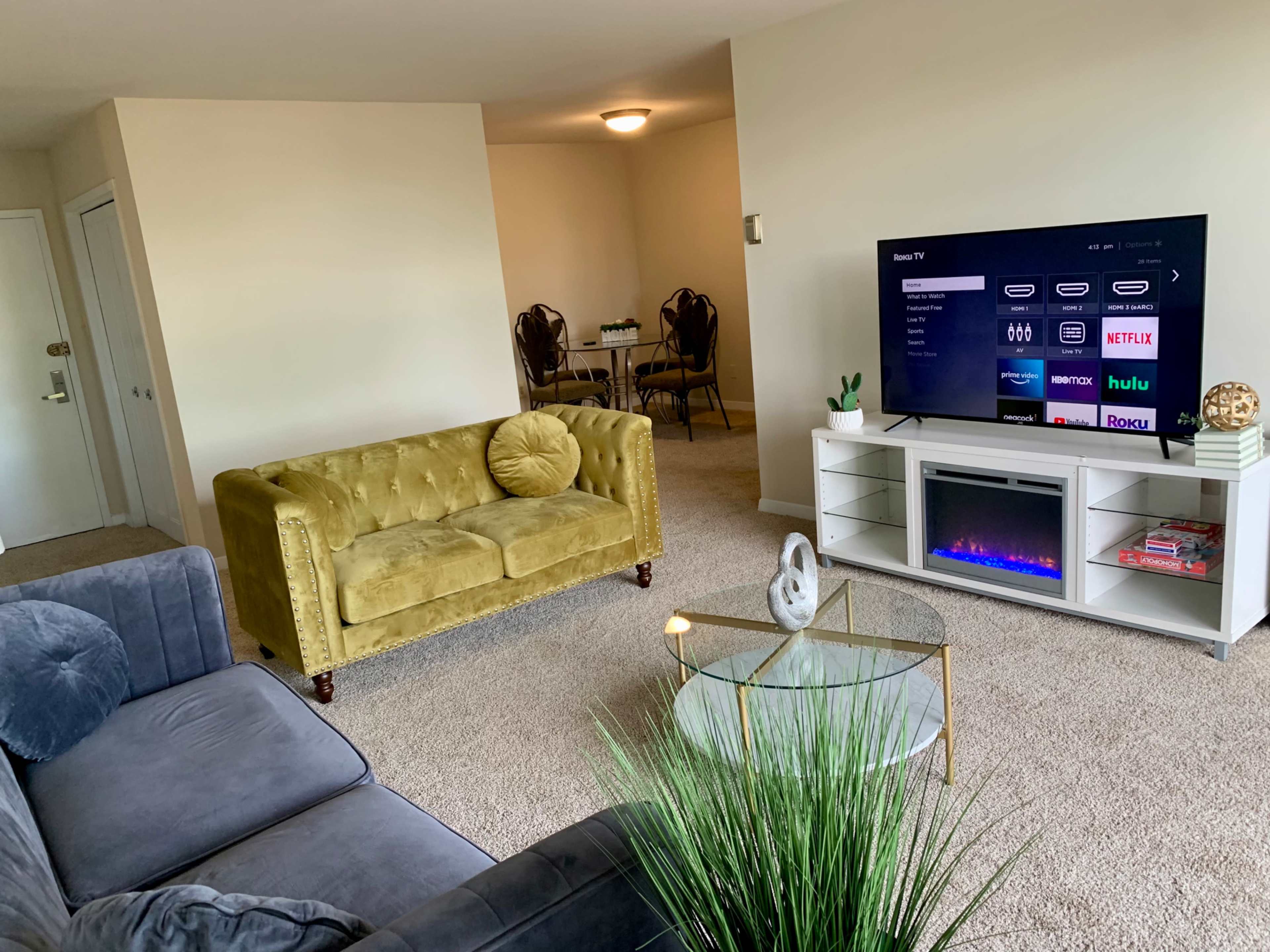 A living room with a green velvet couch, a gray sofa, a glass coffee table, a television displaying streaming options, and a dining area in the background.