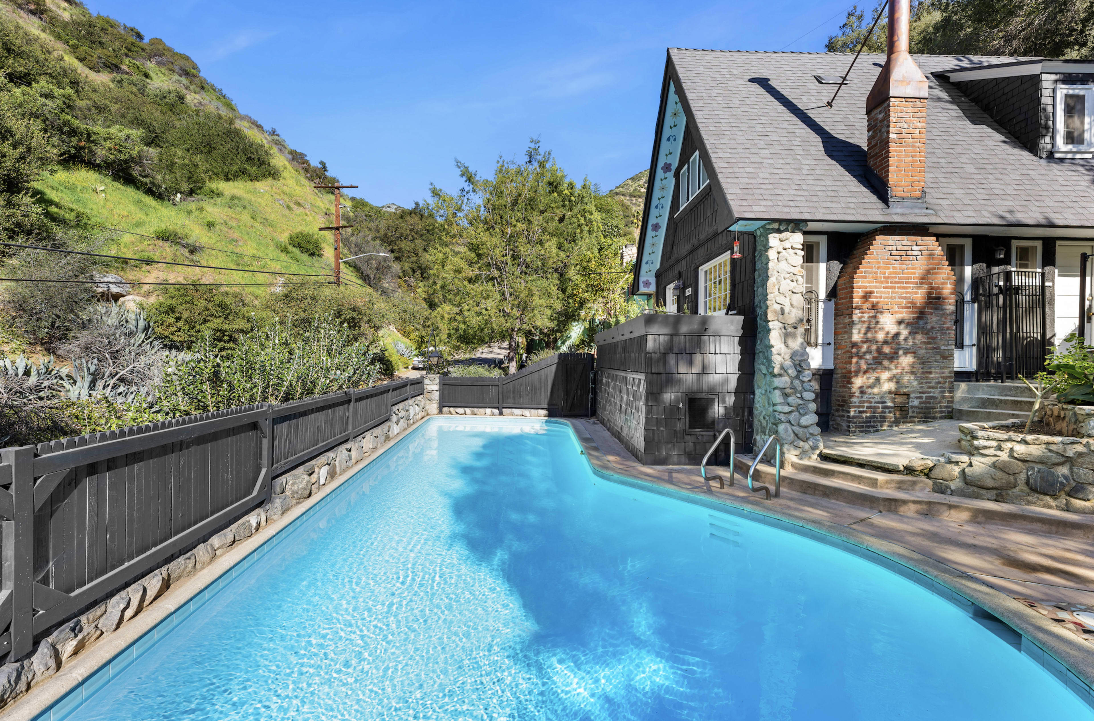 A swimming pool with clear water is situated beside a house, surrounded by greenery and a stone wall.