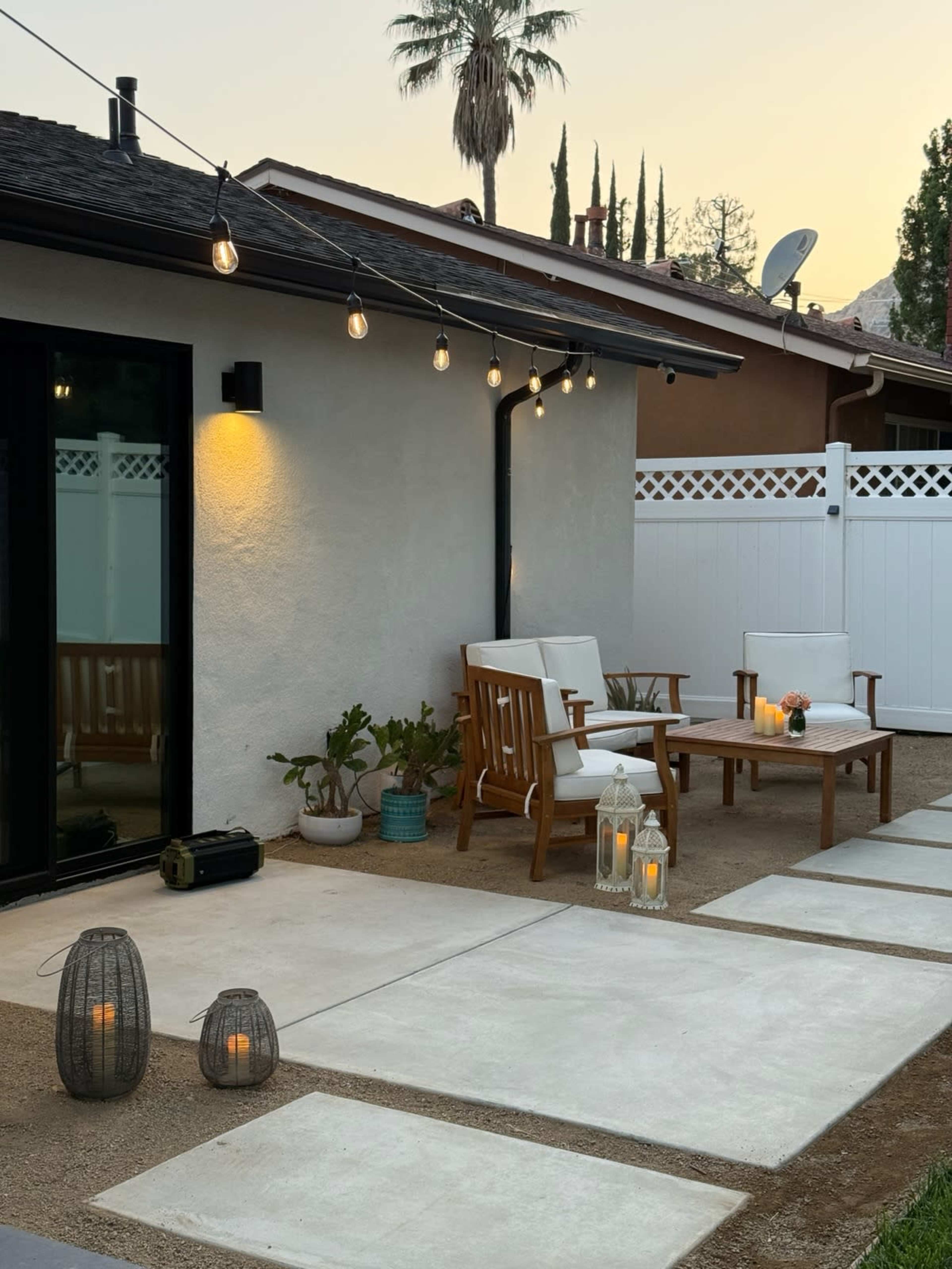 A patio area features two white chairs, a small table with orange flowers, and decorative lanterns situated along a stone pathway under string lights.