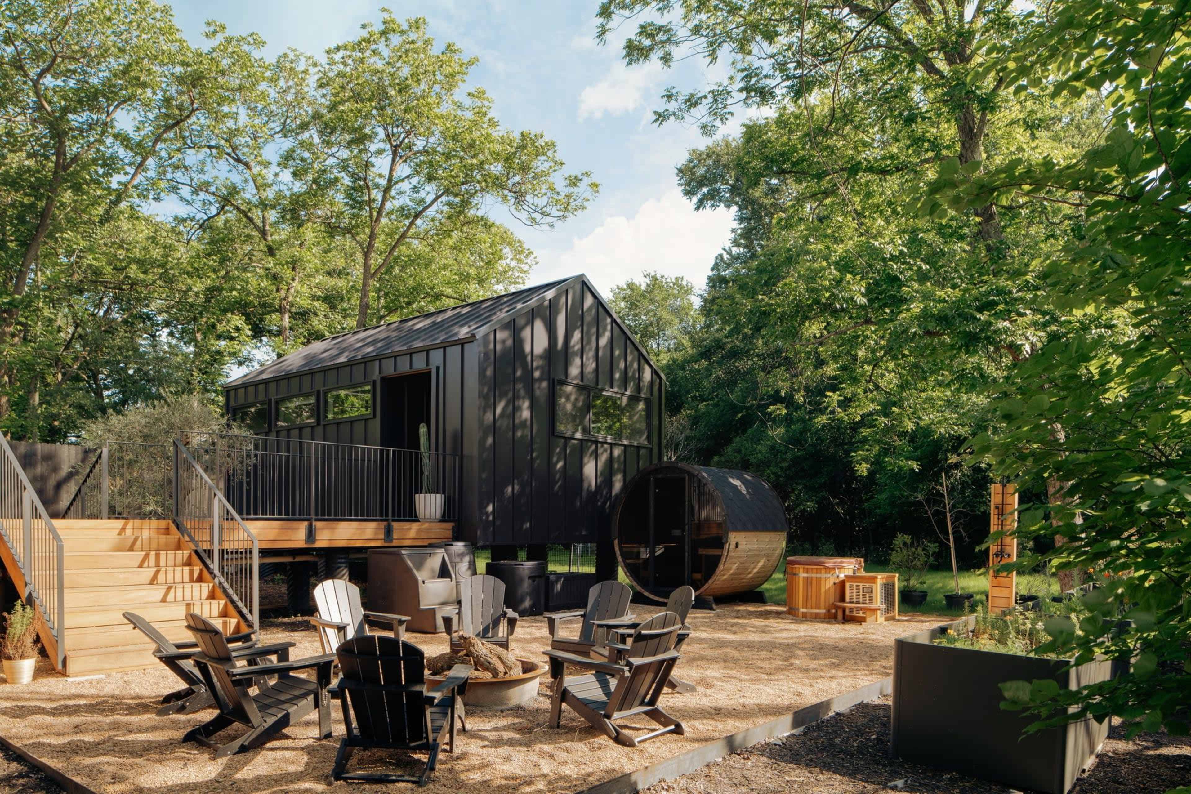A modern black house with a deck overlooks a landscaped yard featuring a circular sauna, outdoor seating, and greenery.