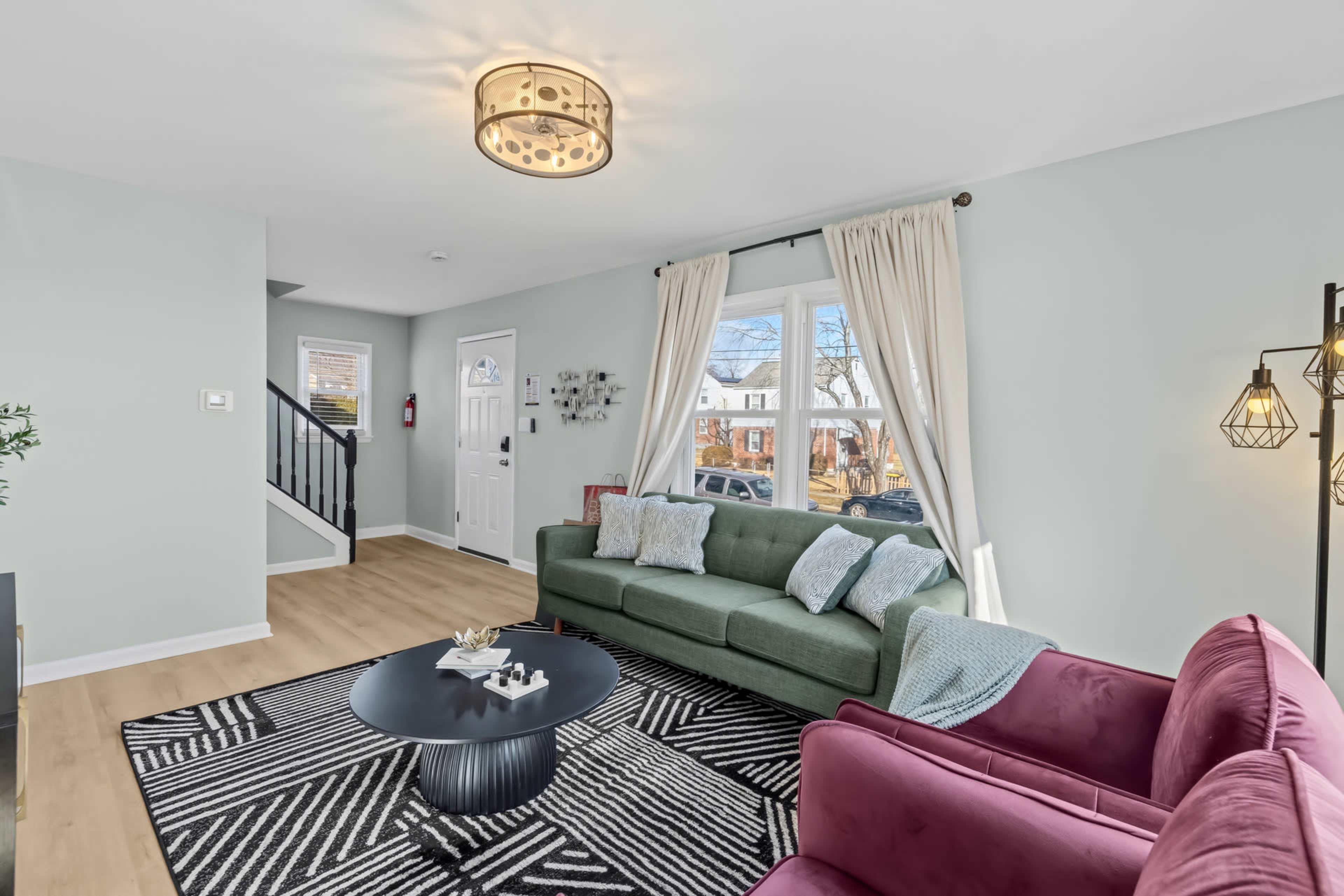 The living room features a green sofa and a pink chair with a patterned rug, large windows, and a staircase in the background.