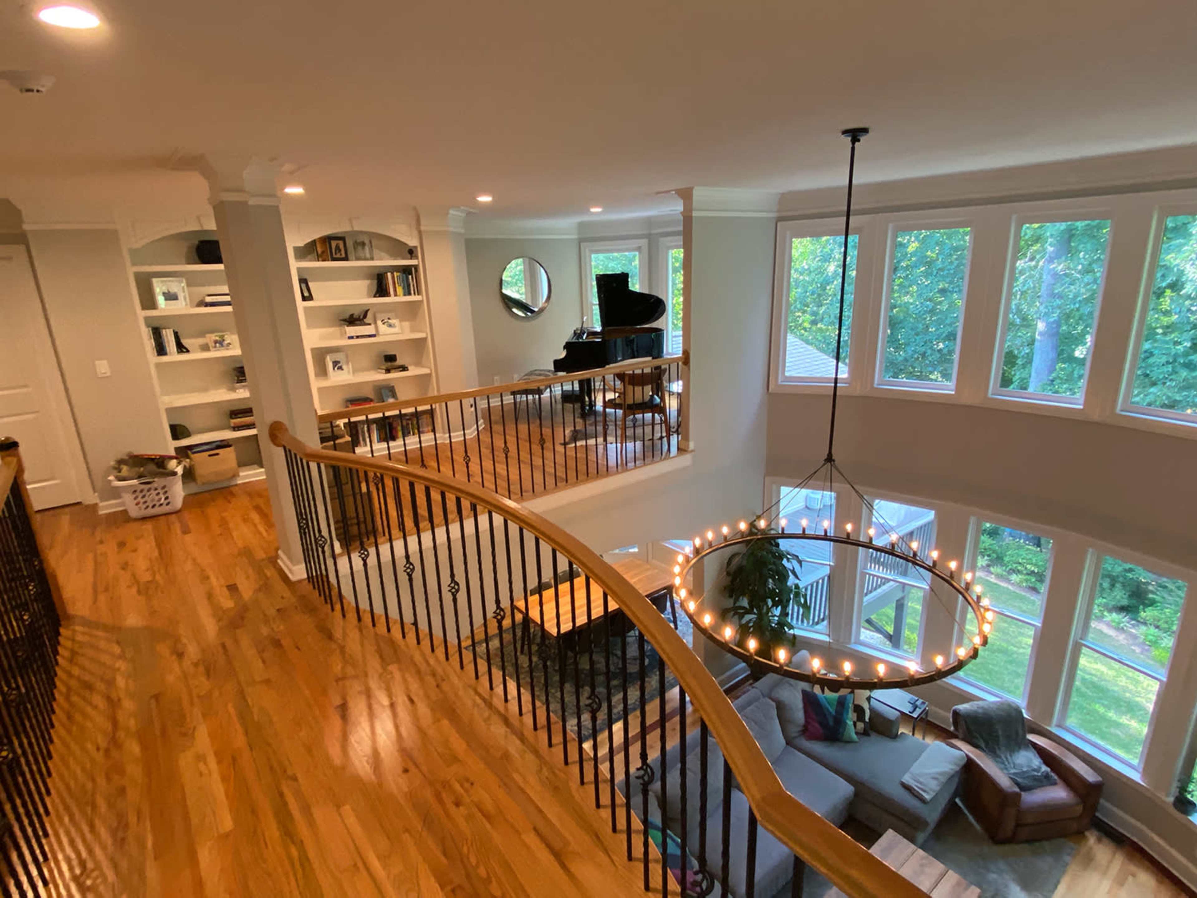 A spacious interior with a wooden staircase, large windows overlooking greenery, a grand piano in the corner, and a modern circular chandelier.