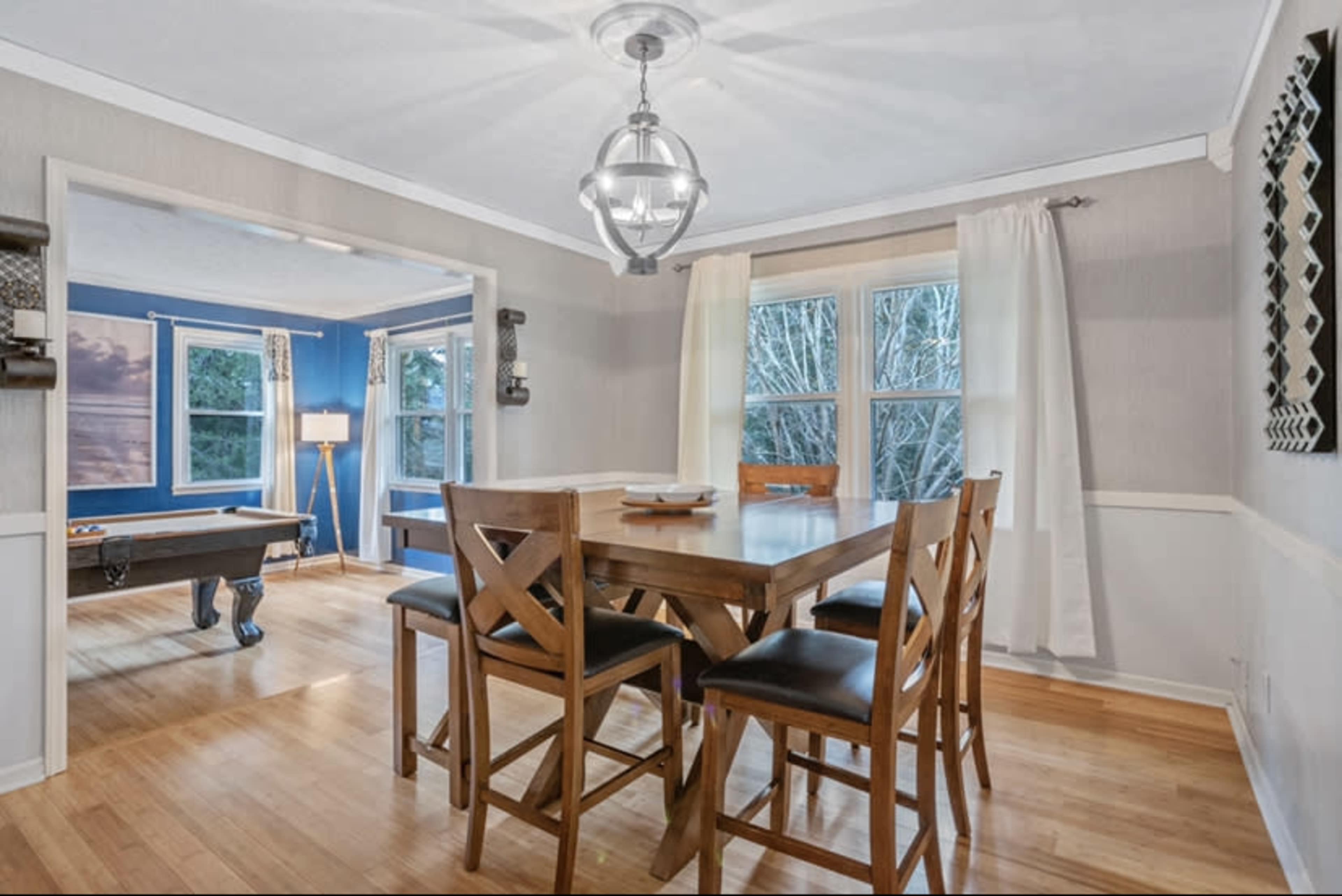 A wooden dining table with high-backed chairs is positioned in a well-lit room that connects to a blue-walled space with a pool table.