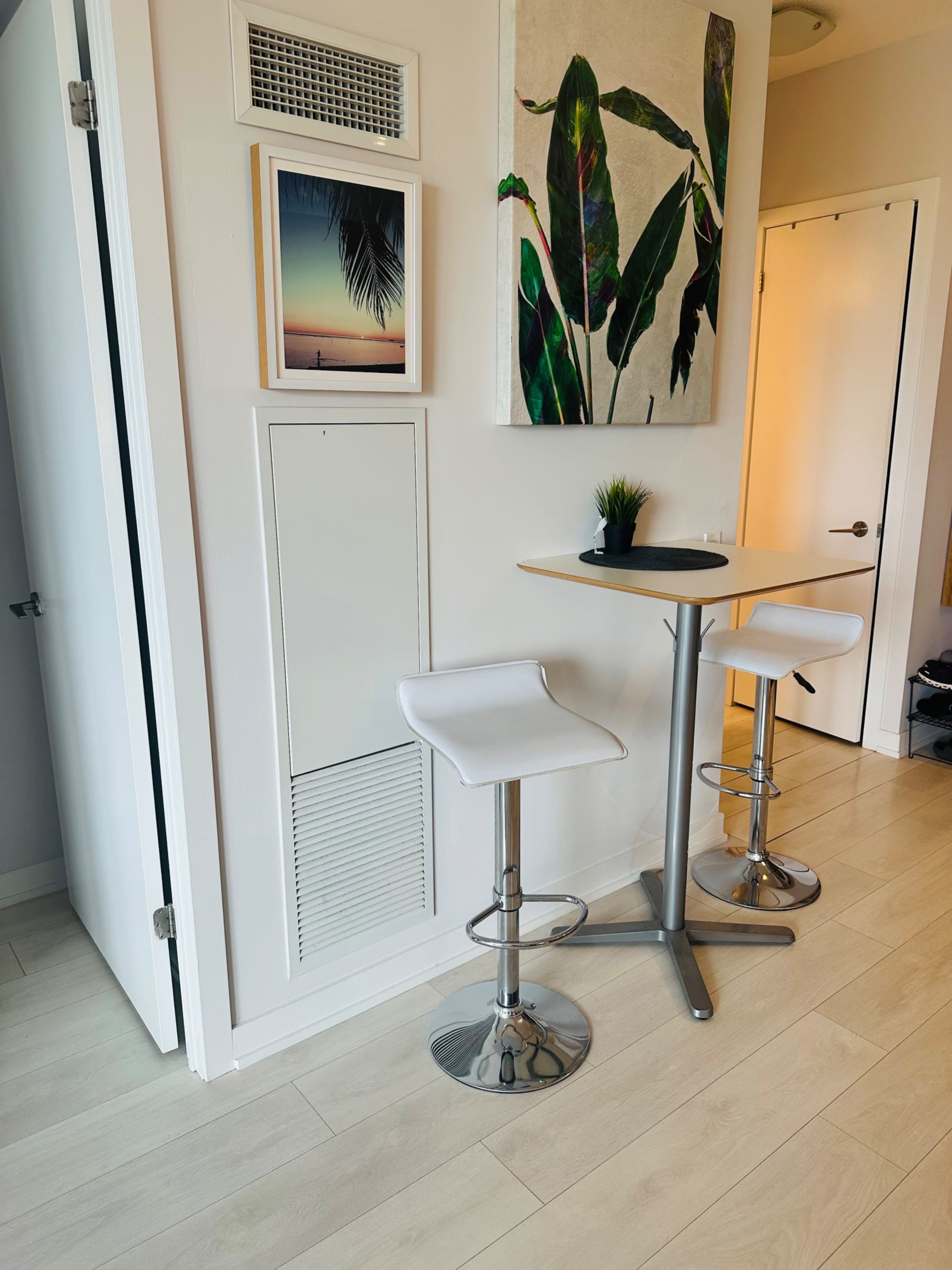 A small dining area features two white bar stools and a round table against a wall adorned with framed artwork and a potted plant.