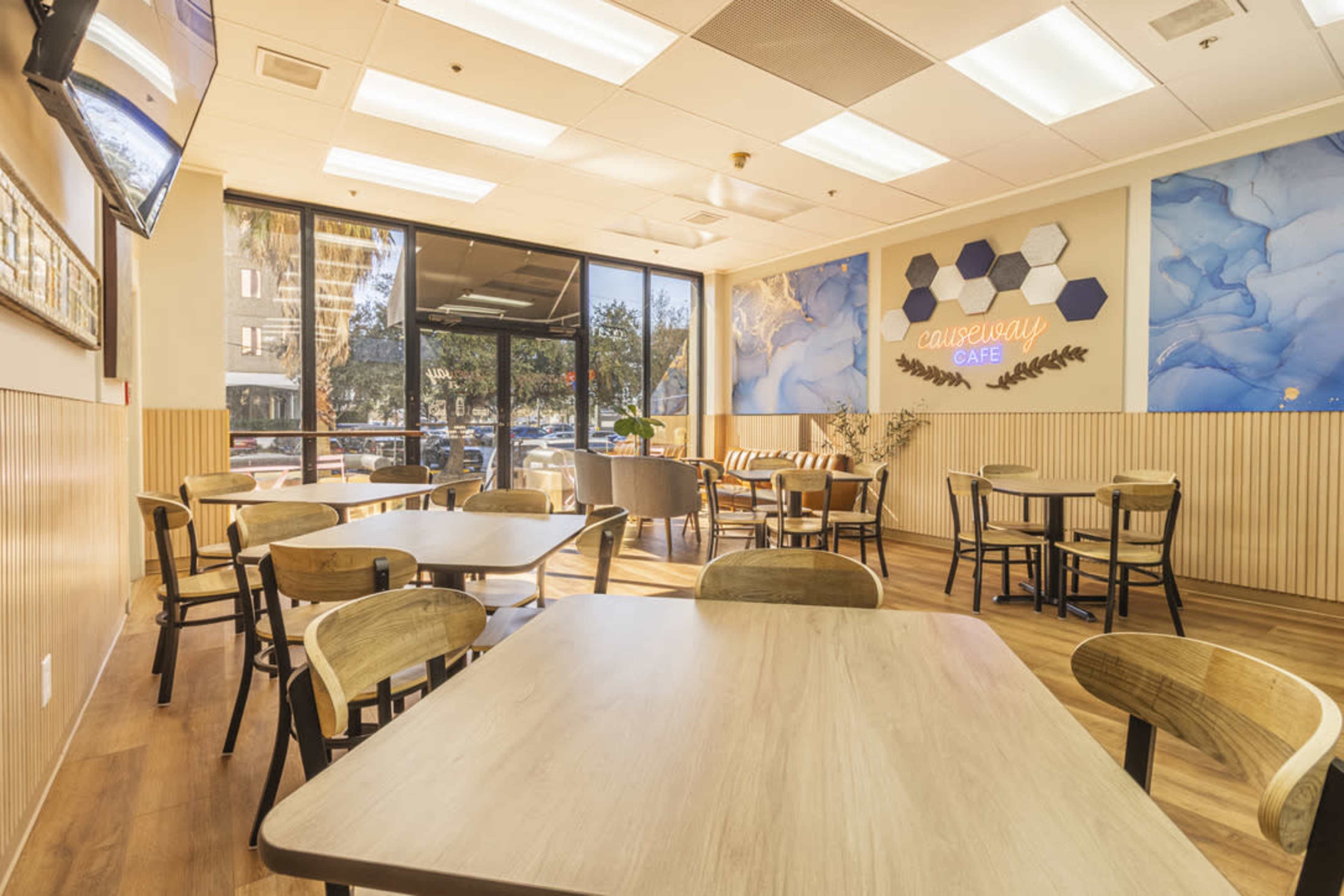 The image shows a modern café with wooden tables and chairs, large windows letting in natural light, and decorative wall art featuring hexagonal shapes.