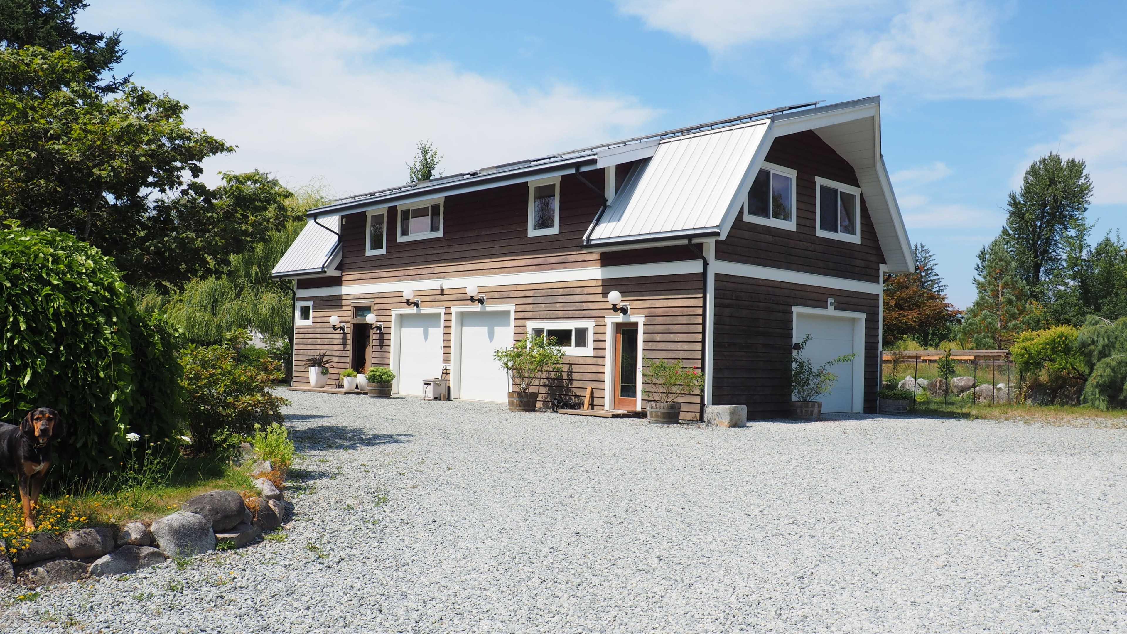 A two-story wooden building with a sloped metal roof is set on a gravel driveway surrounded by greenery.
