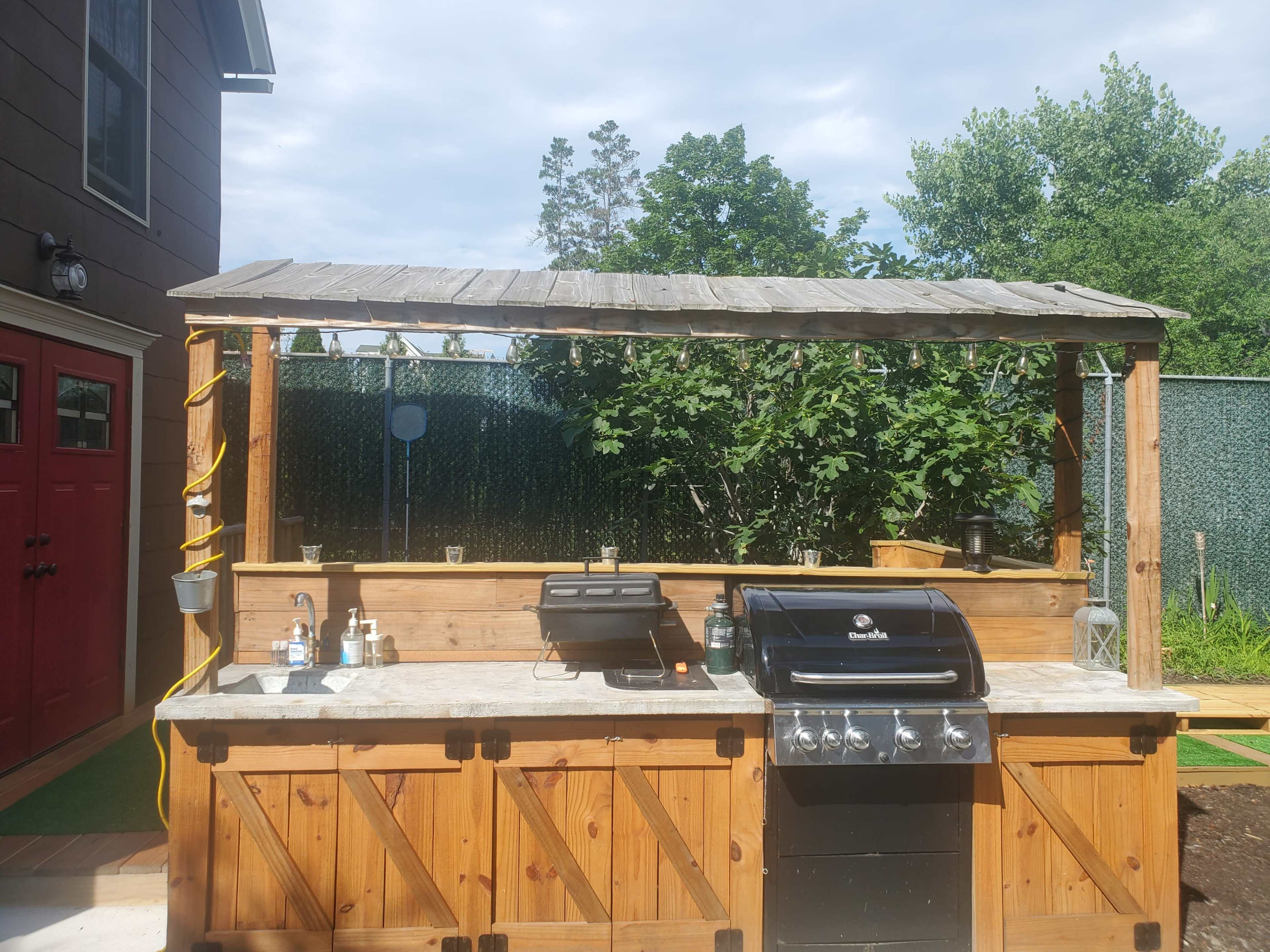 An outdoor kitchen with a stone countertop, a gas grill, and a shaded area under a wooden structure.