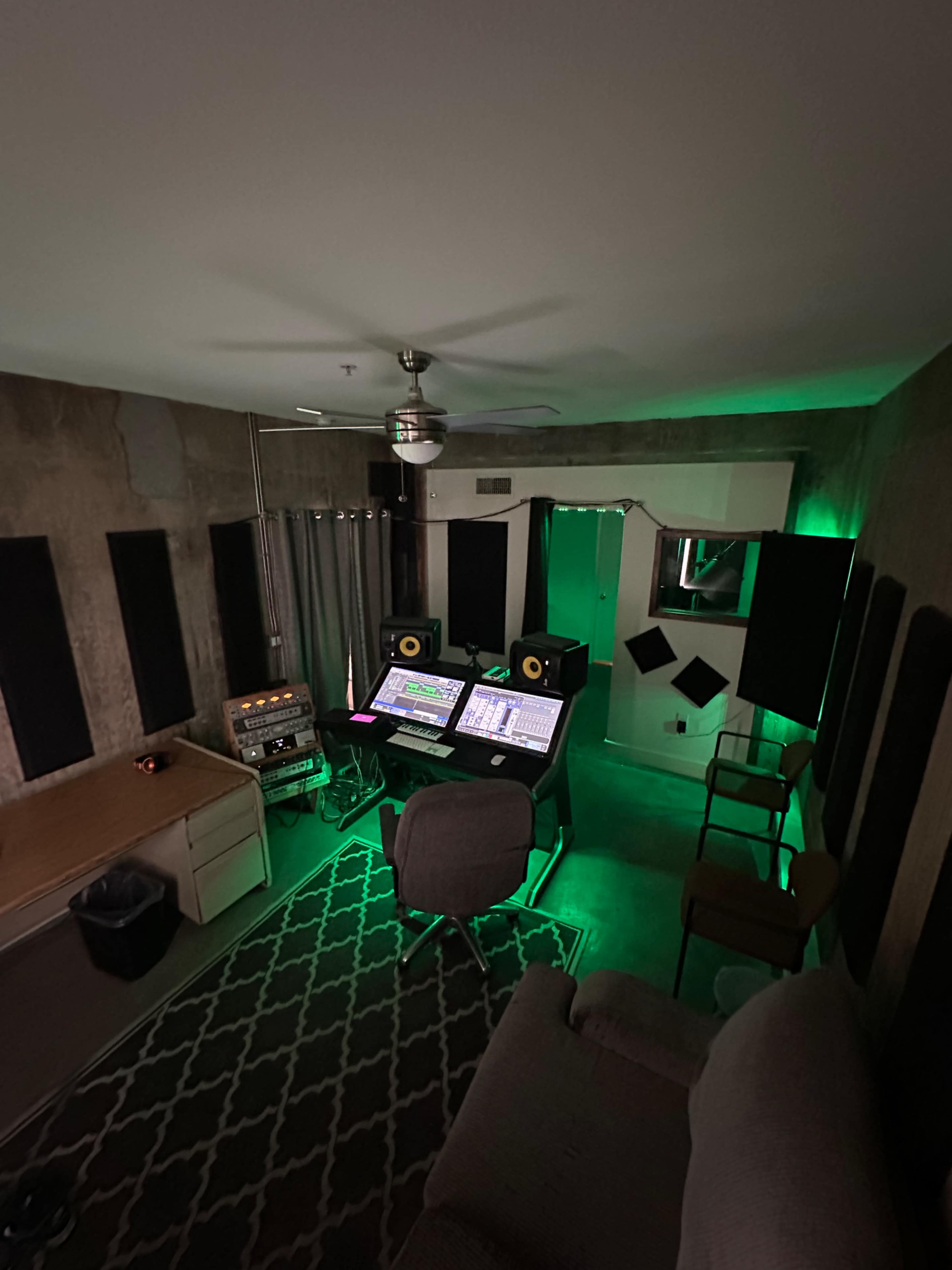 A small, dimly lit music studio featuring a desk with dual computer monitors, audio equipment, and green ambient lighting.
