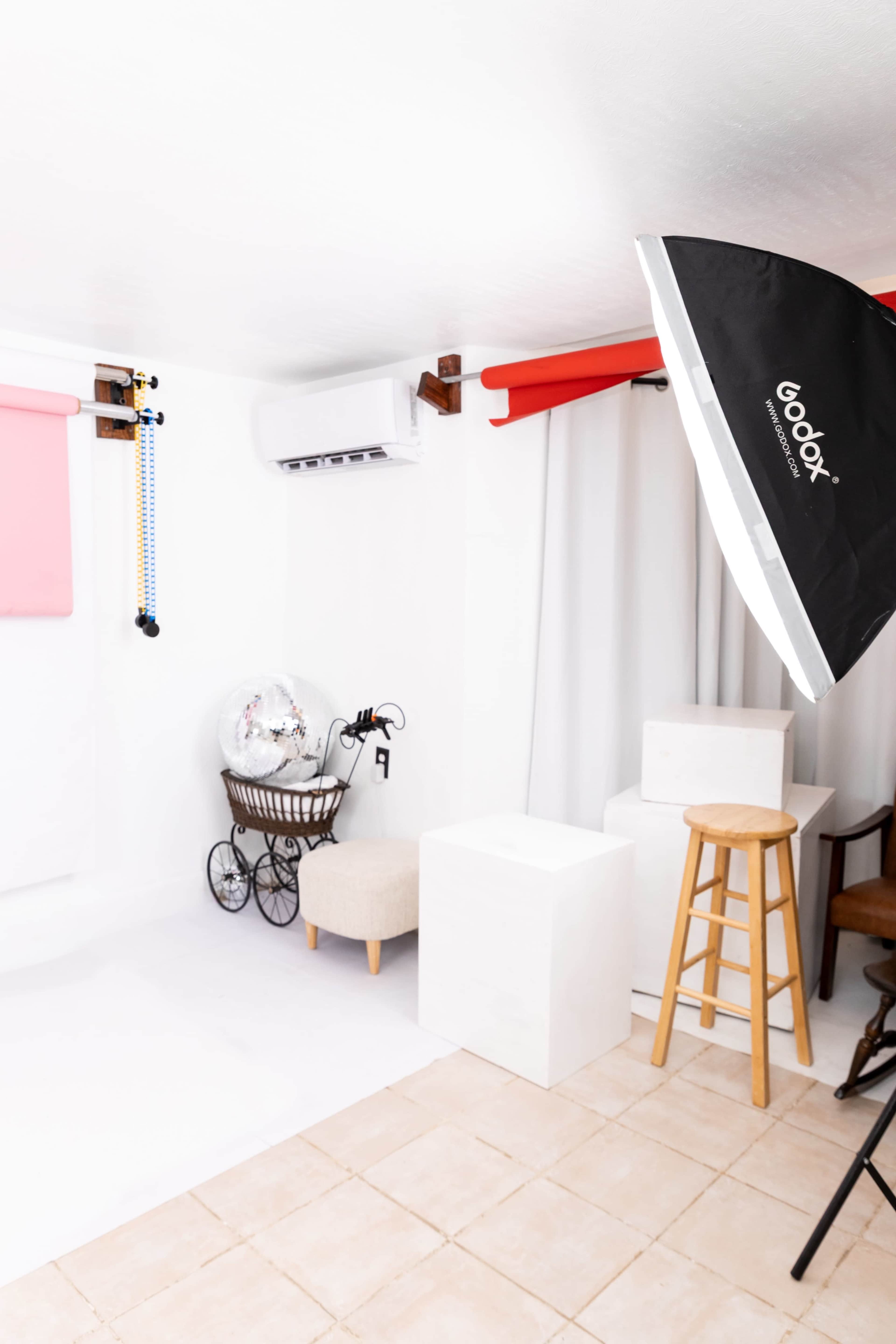 The image shows a photography studio with a white backdrop, various props, a light stand, and a chair arranged in the space.