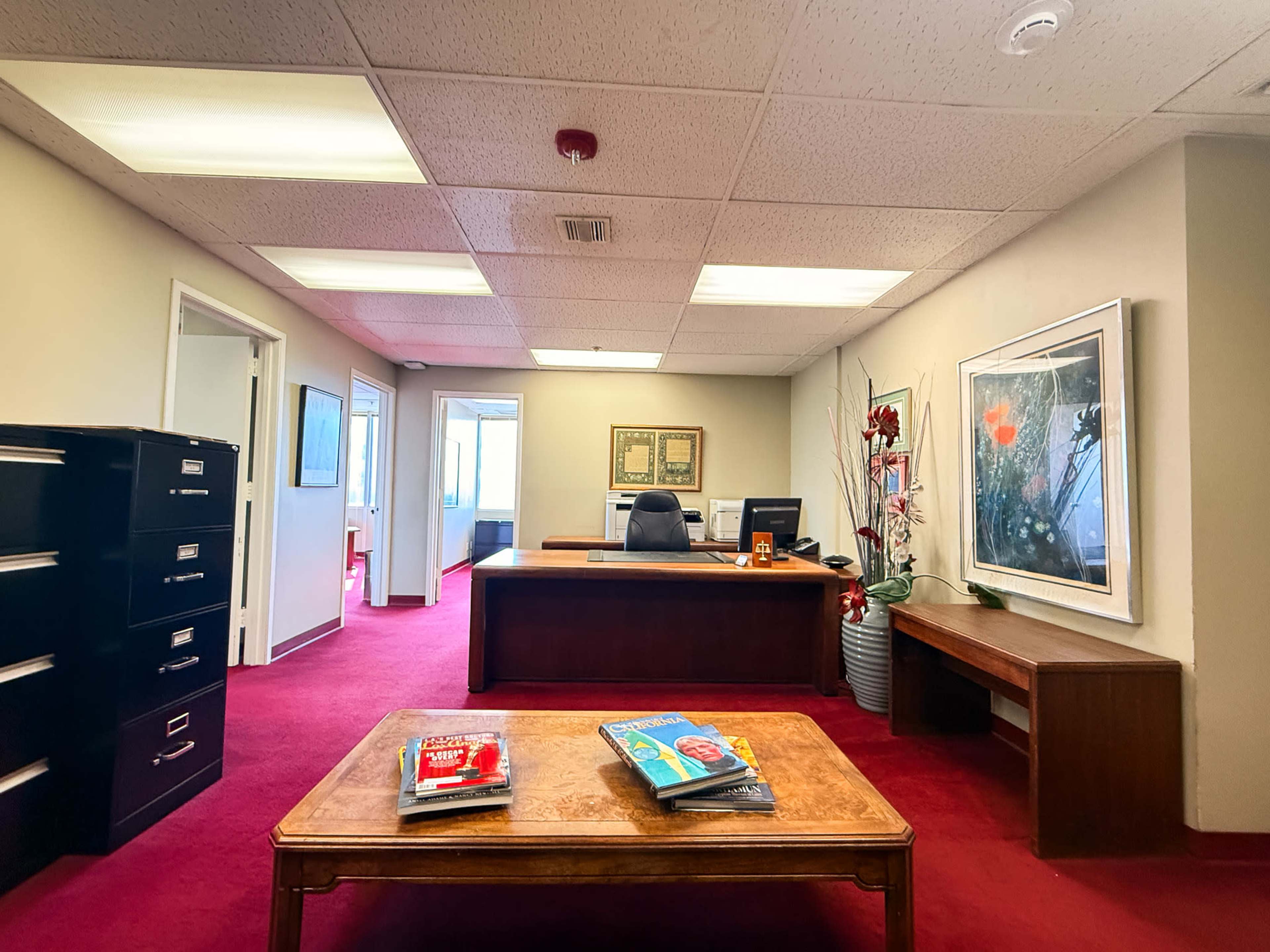 The image shows an office space featuring a desk with a chair, a coffee table with magazines, filing cabinets, and artwork on the walls.