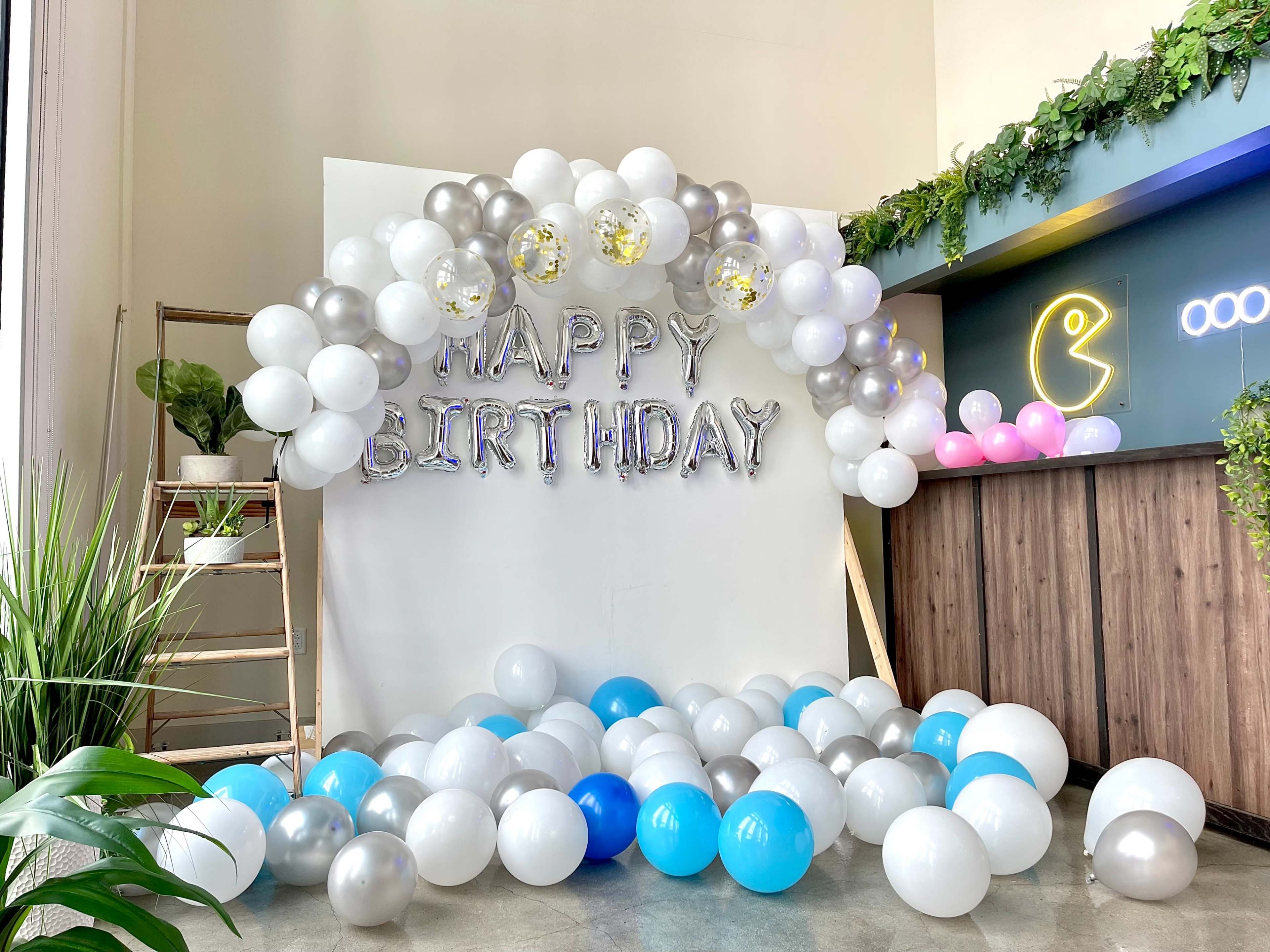 A birthday decoration setup with a "HAPPY BIRTHDAY" banner beneath a balloon arch, surrounded by a mix of blue, silver, and white balloons on the floor.