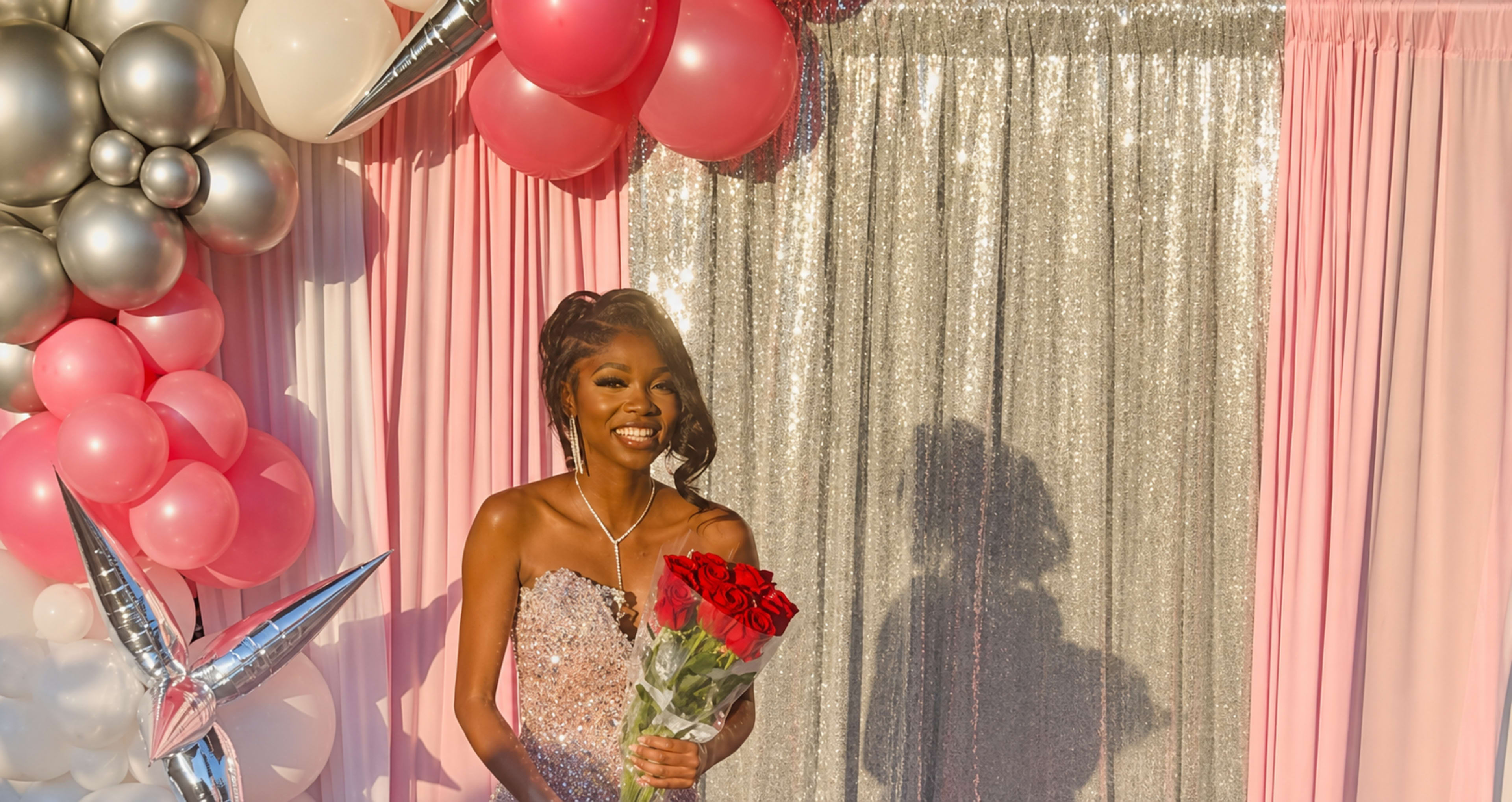 A woman in a sparkling dress holds a bouquet of red roses in front of a backdrop of pink and silver balloons and a sequined curtain.