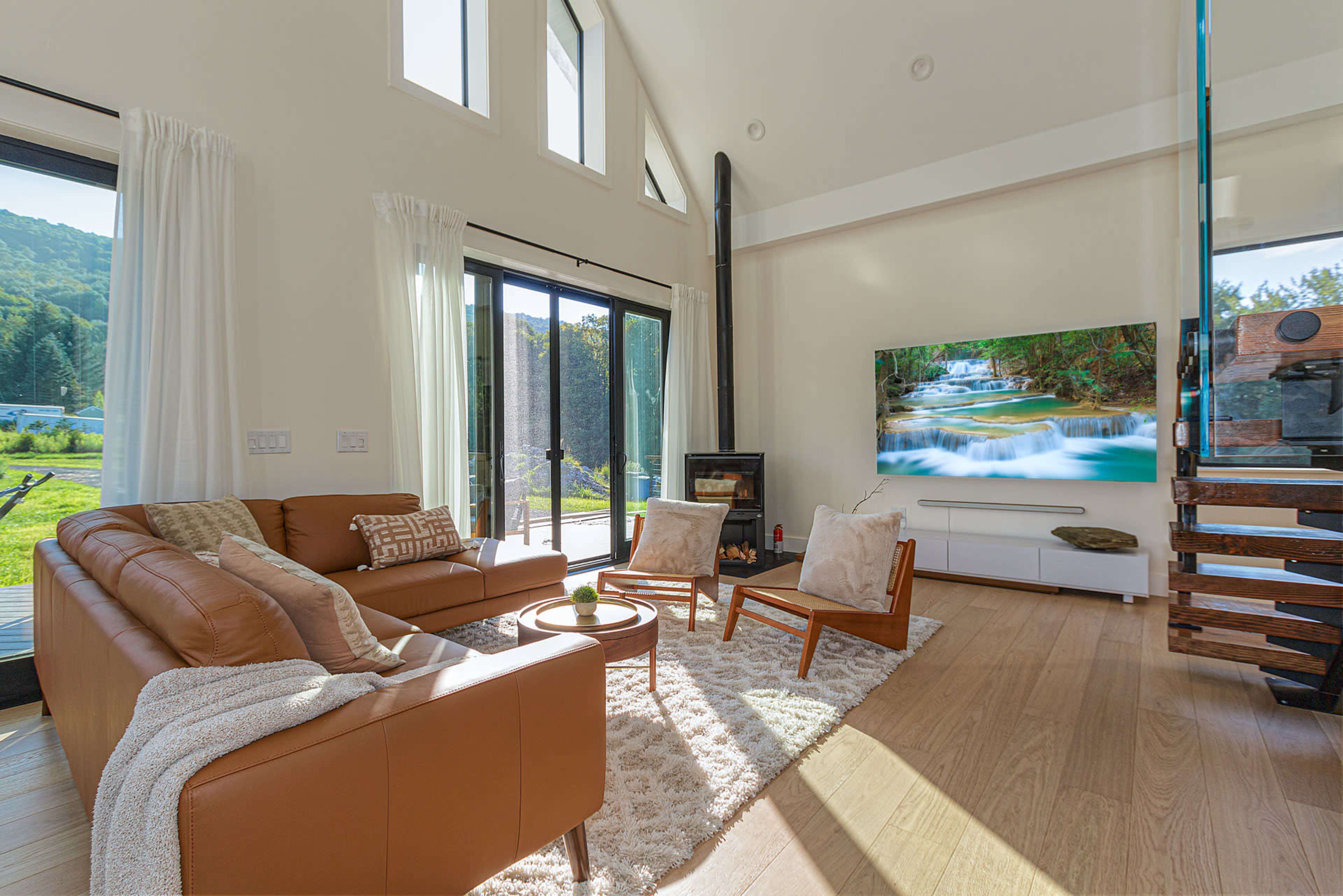 A modern living room with a leather sofa, two chairs, and a large TV displaying a nature scene, surrounded by floor-to-ceiling windows.