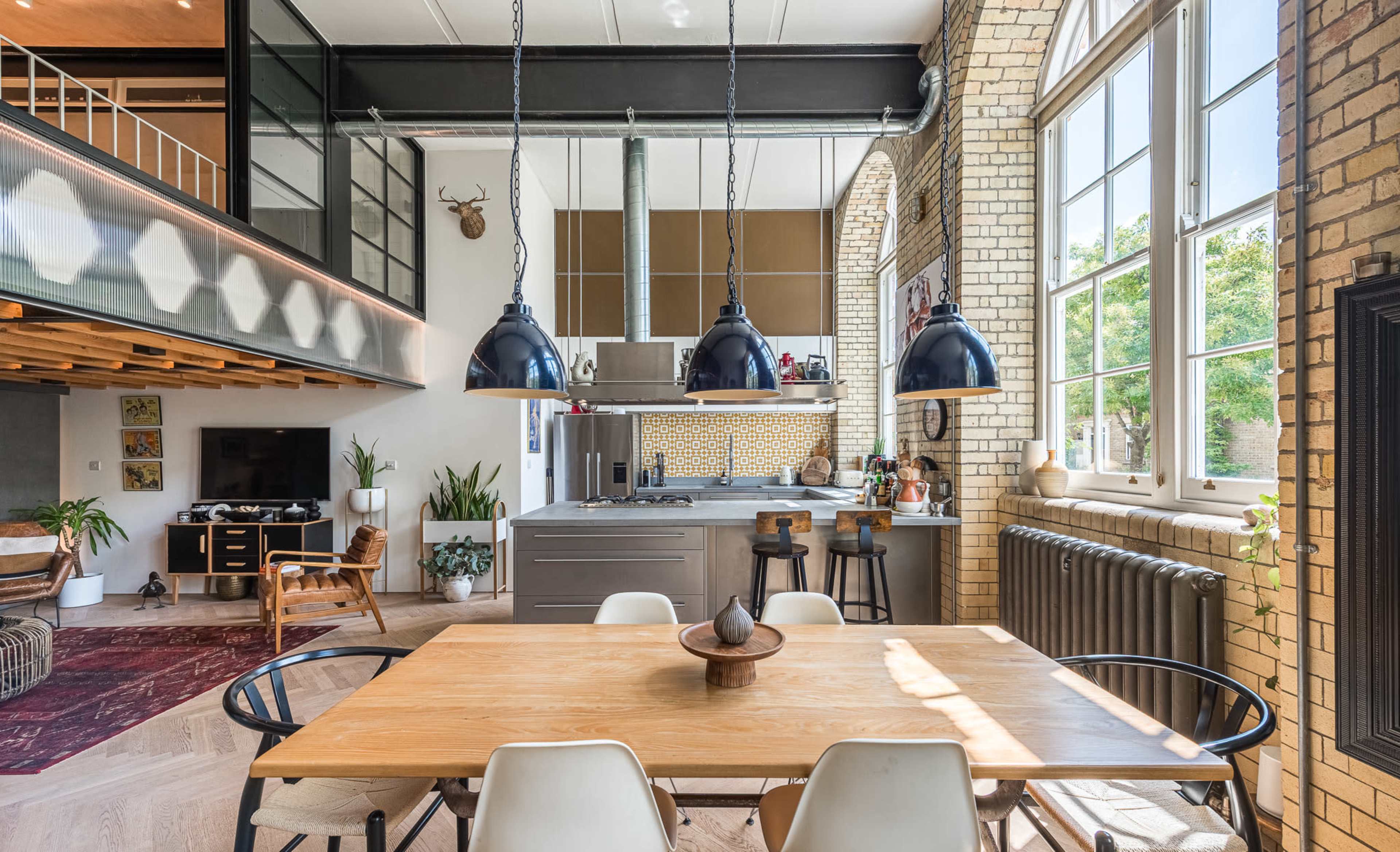 The image shows a modern kitchen and dining area with a wooden table, black pendant lights, and large windows allowing natural light to fill the space.