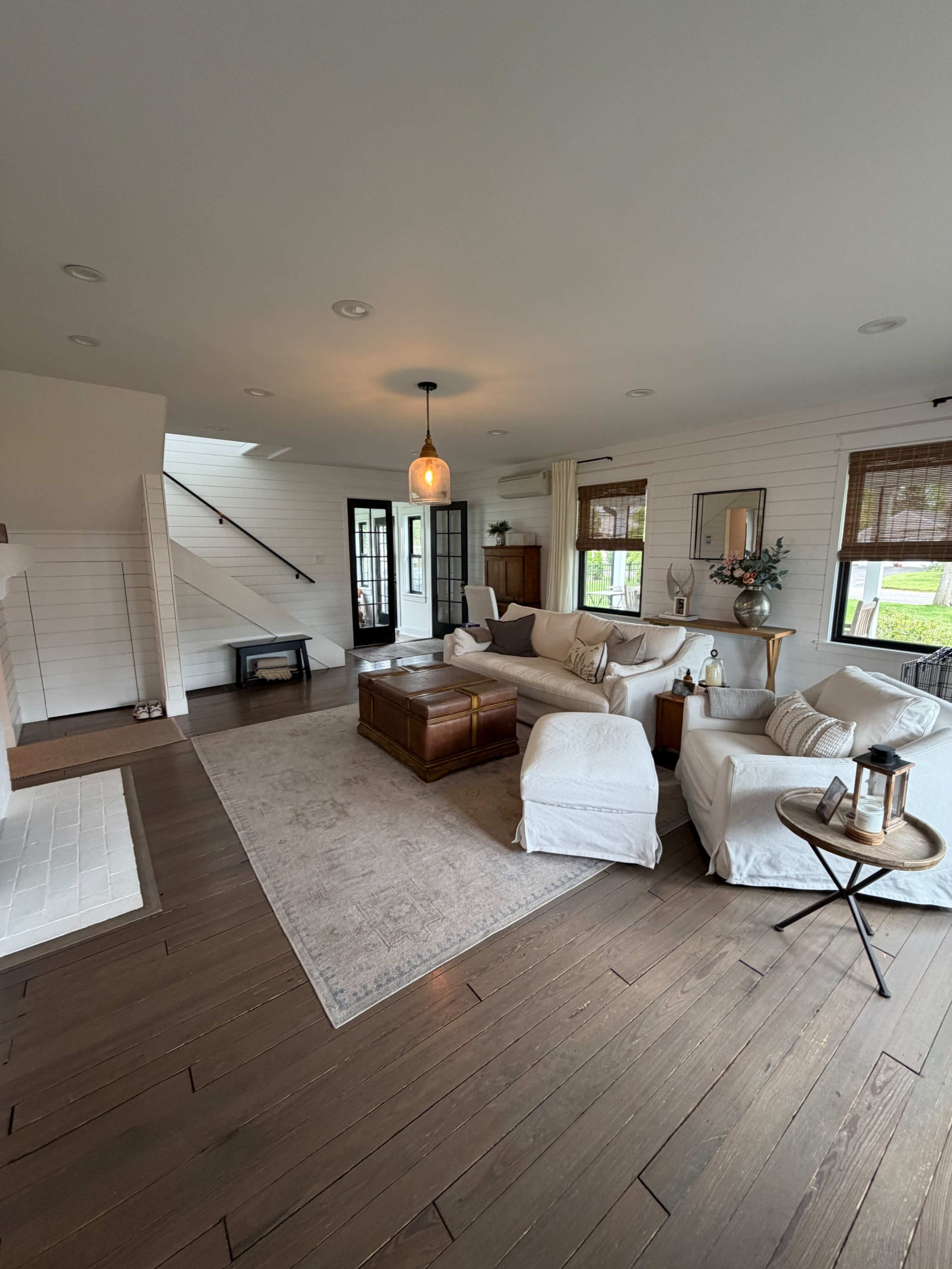 The image shows a modern living room with light-colored furniture, a wooden coffee table, and a staircase leading to another level.