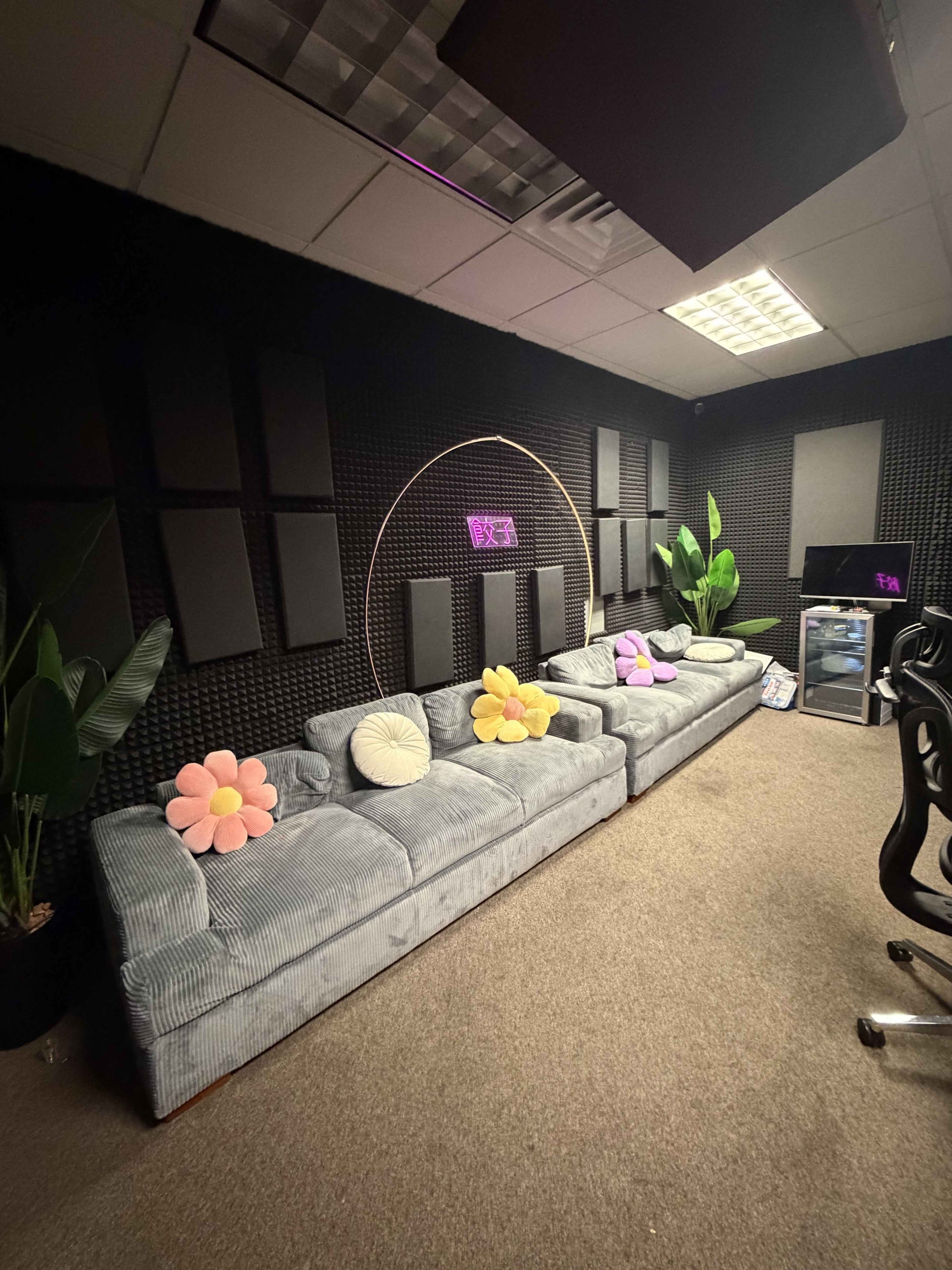 The image depicts a lounge area featuring a gray striped sofa adorned with colorful flower cushions, surrounded by green plants and soundproofing panels on the walls.
