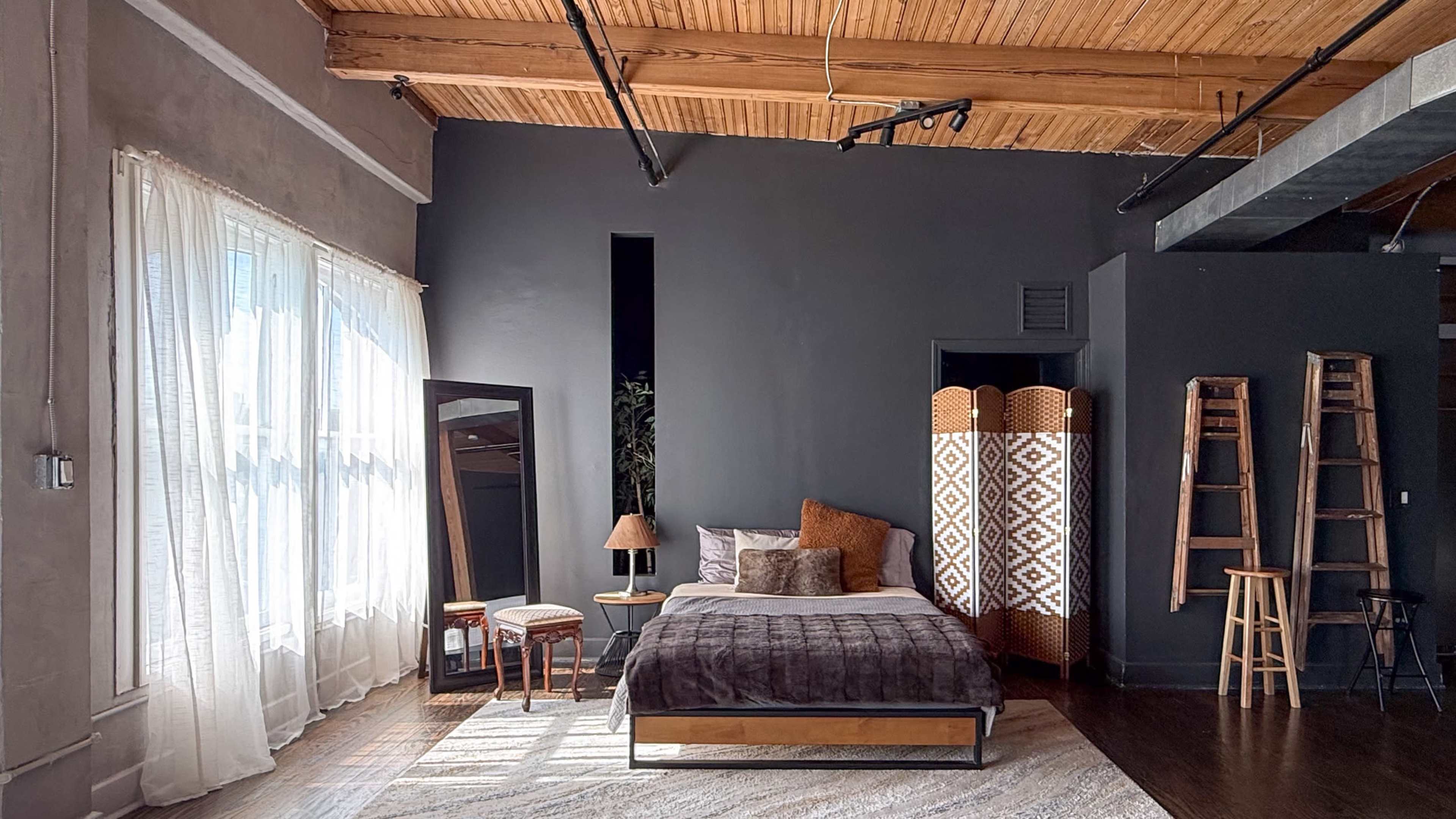 The image features a modern bedroom with a large bed, a mirror, and a decorative folding screen, illuminated by natural light from a large window.