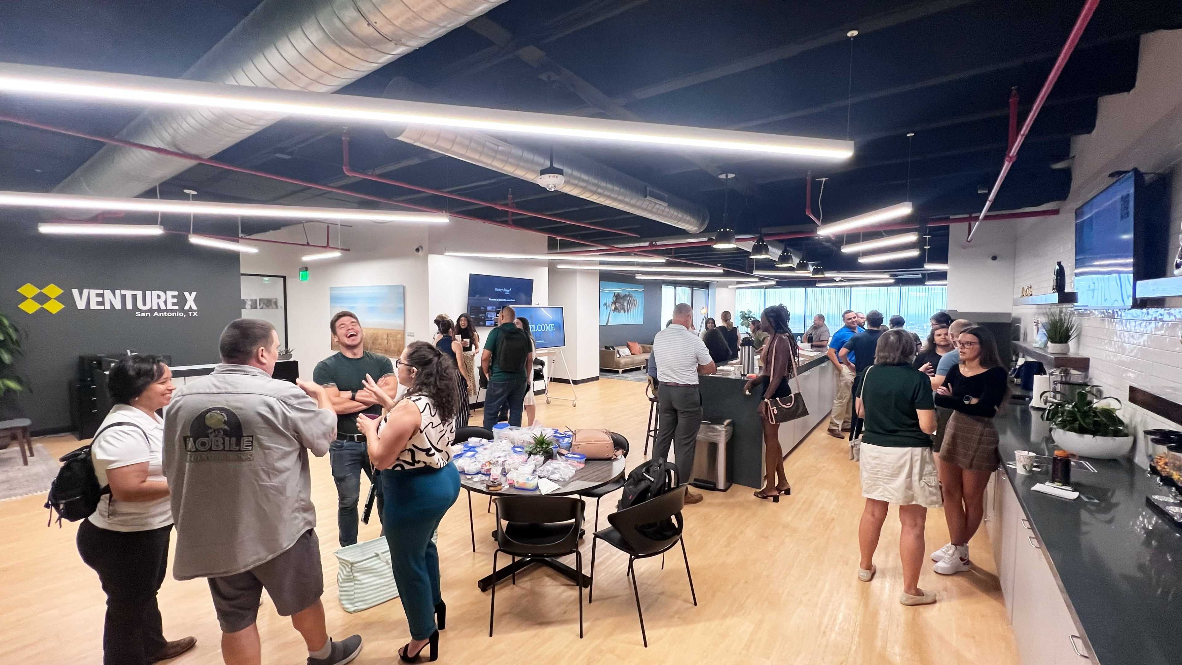 A group of people are networking at a business event in a modern office space with seating areas and a refreshment table.