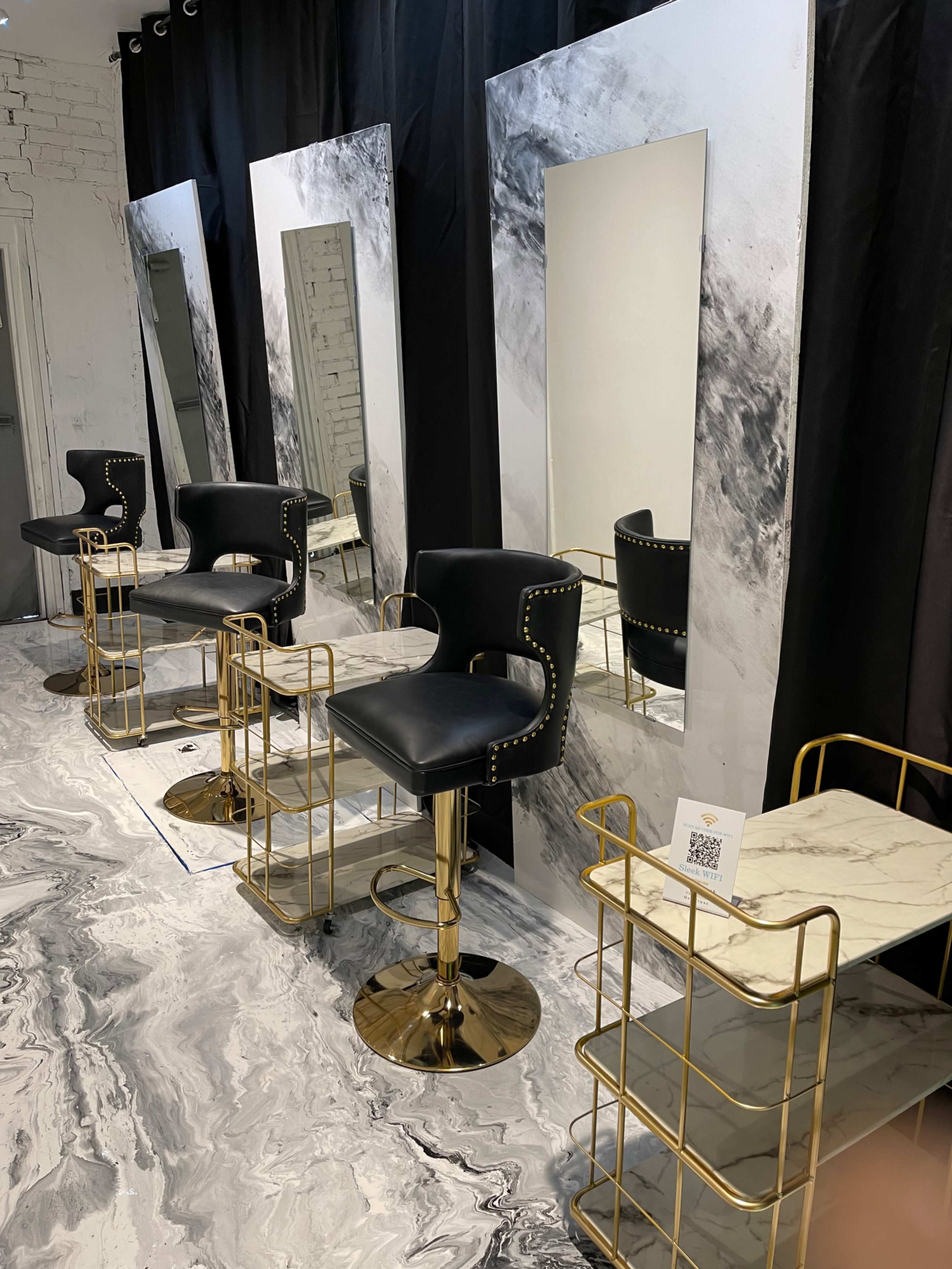 A modern salon interior with black and gold bar stools, large mirrors, and a marble-patterned floor.